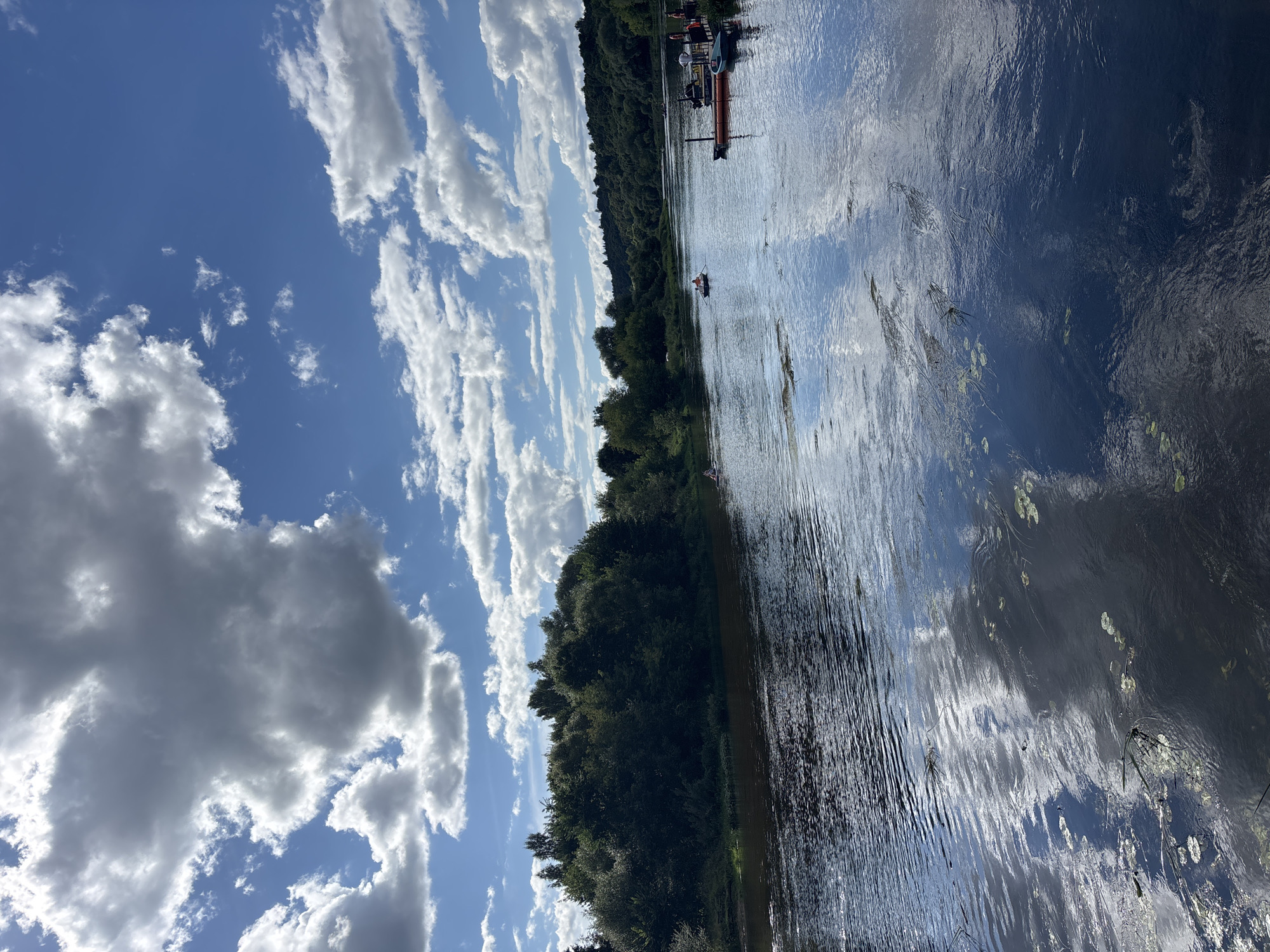 The image depicts a serene riverside scene under a dynamic sky. The Moskva River flows through a lush green landscape, with dense trees lining the banks. The water reflects the bright blue sky and the scattered, cumulus clouds, creating a mirrored effect. Several small boats are visible on the water, some with people in them, suggesting recreational activity. On the right bank, a wooden dock with several moored boats is present, along with a few individuals. The atmosphere is peaceful and inviting, hinting at a pleasant summer day. This scene is likely located along the Moskva River in Zvenigorod, Russia, a town known for its natural beauty and proximity to the river.