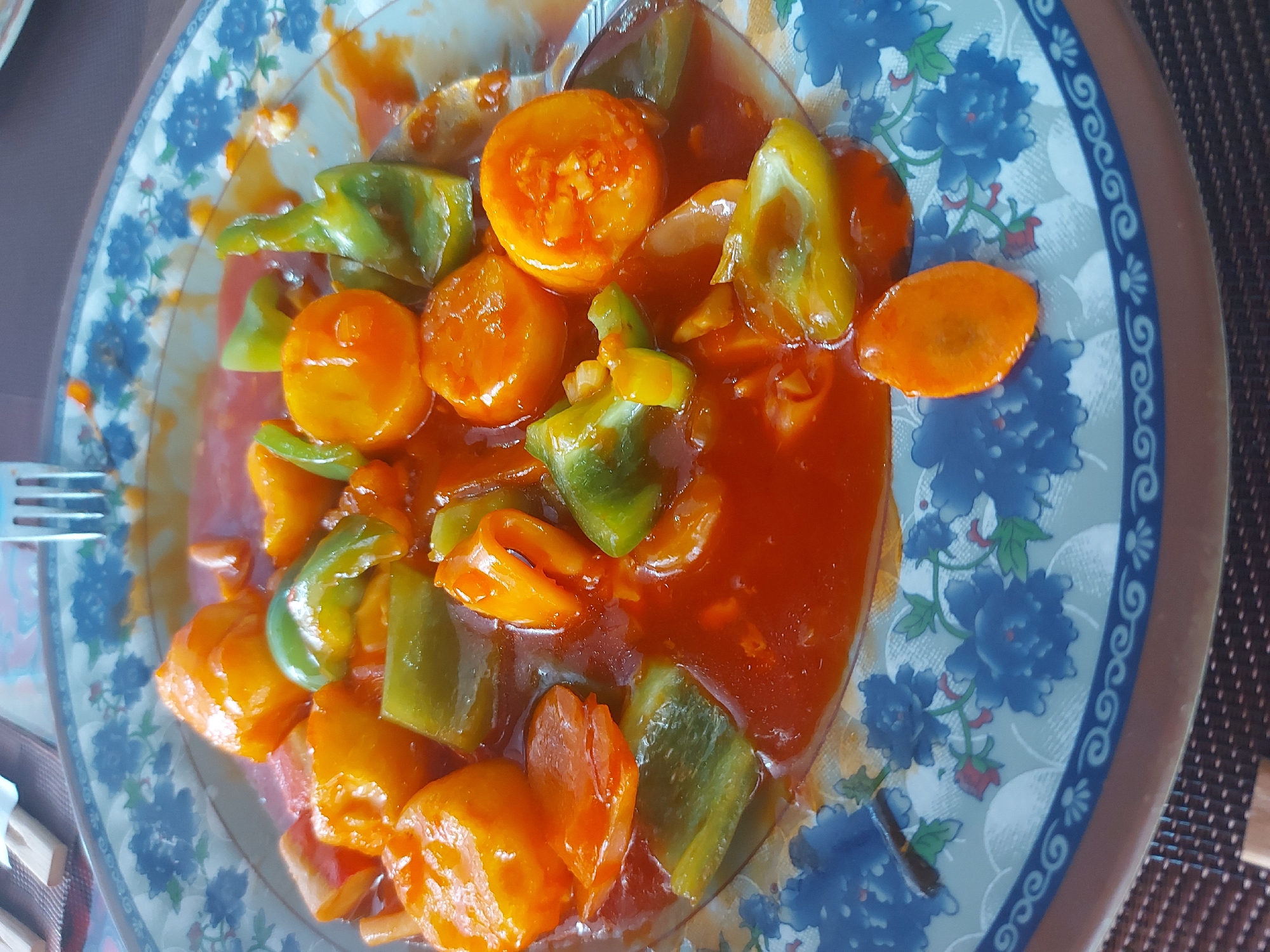 A close-up, overhead shot shows a plate of sweet and sour chicken with bell peppers and carrots, coated in a vibrant red sauce. The dish is served on a plate with a blue floral pattern around the rim. A fork is partially visible on the right side of the plate. The plate rests on a brown textured placemat, and in the background, wooden chopsticks and the edge of another plate are visible. The overall lighting is bright, suggesting an indoor setting, possibly a restaurant in Blagoveshchensk, Russia.