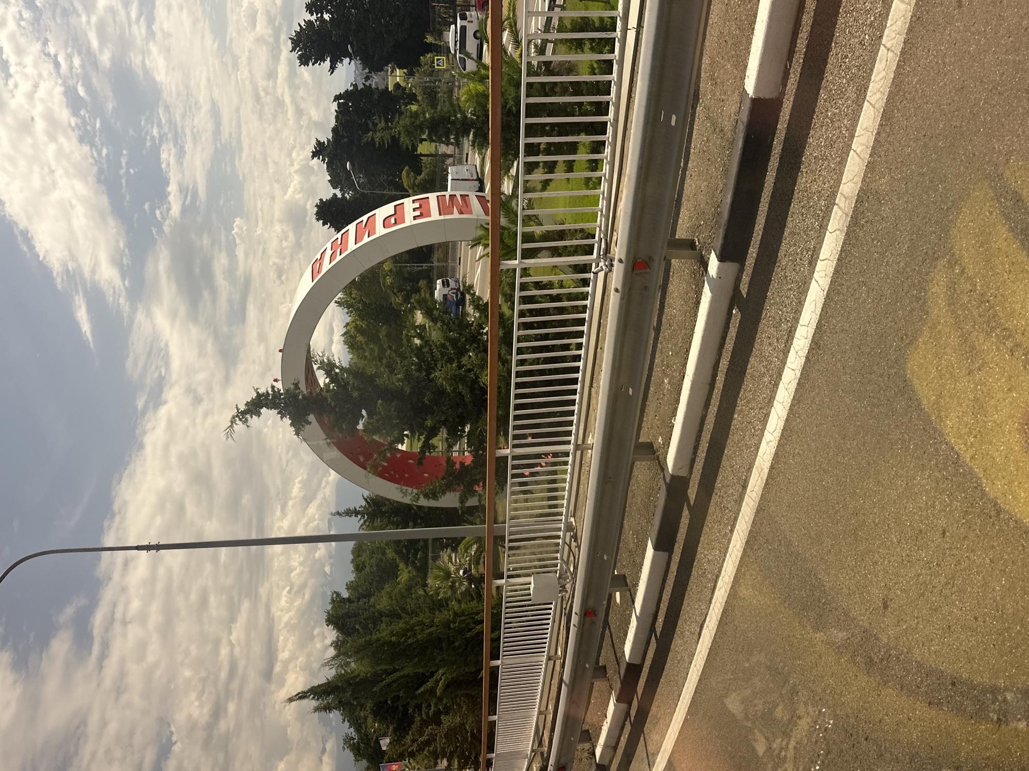 This is a photograph of an arched structure with red lettering, likely a gateway or monument, in Sochi, Russia. The lettering on the arch appears to spell out "МЕРЧАНТ" (Merchant). The arch is white with red accents. It is surrounded by trees and vegetation. In the background, there are roads with vehicles, including vans and a car. A street light is visible to the left of the arch. The sky is partly cloudy. The foreground shows a road with yellow and white markings, and a metal guardrail and white fence. The overall scene suggests an outdoor environment, possibly a park or a city entrance.