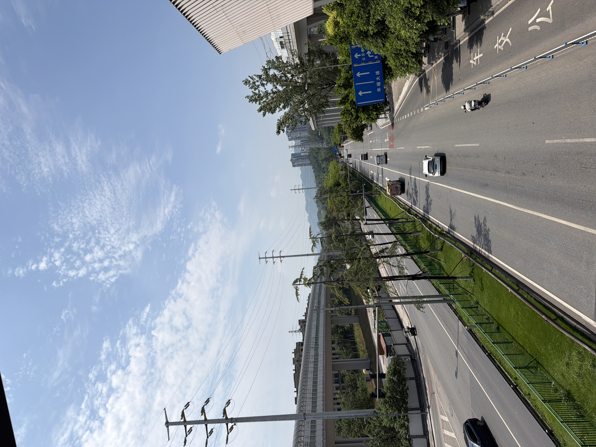 This is a high-angle, full shot of a multi-lane highway in Tiaodeng, China, on a clear day with scattered white clouds. The highway stretches into the distance, curving slightly to the right. On the left side of the highway, there is an elevated pedestrian walkway with a translucent roof, supported by concrete pillars. Beyond the walkway, there are buildings and trees. On the right side of the highway, there are more trees and a modern, beige building with a slatted facade. Traffic is present on the highway, including cars, a truck, and a motorcycle. The road is marked with white lines. A blue traffic sign with white arrows and Chinese characters is visible on the right side of the road. Overhead power lines and utility poles are also visible. The overall scene depicts a contemporary urban environment with infrastructure and daily activities.