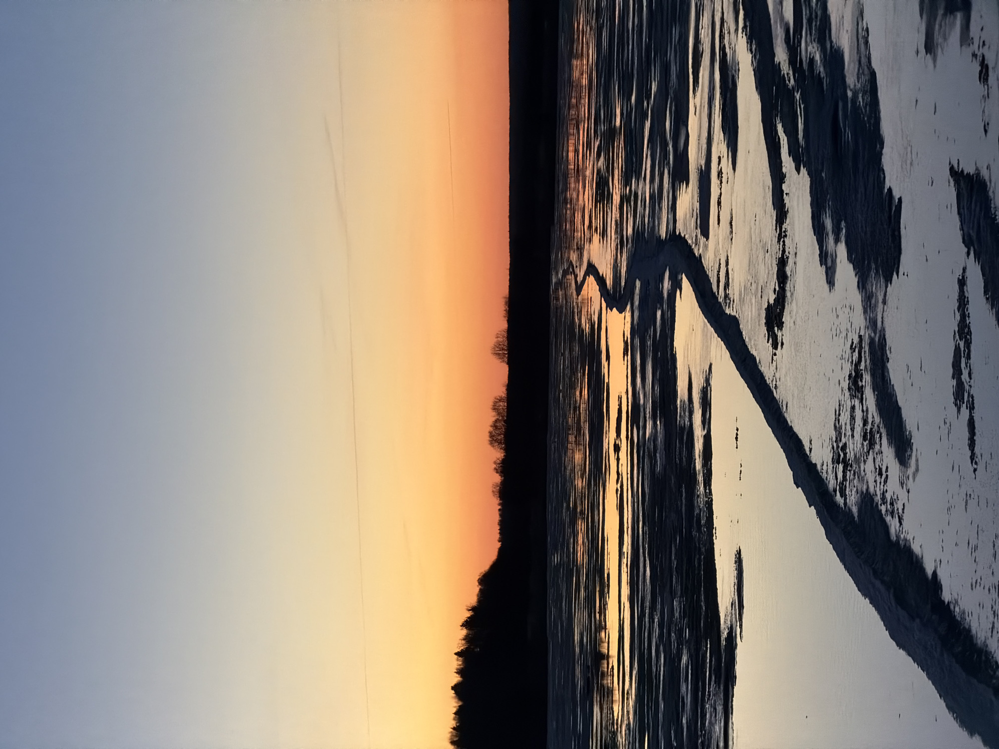 The image depicts a winter scene at sunset, possibly on the Izhevsk Pond (Izhevskiy Prud) in Izhevsk, Russia. The sky transitions from blue to orange-yellow near the horizon. A dark line of trees and bushes is visible along the horizon line. The foreground shows a partially frozen body of water, likely the pond. The ice exhibits patterns of dark and light areas, potentially indicating varying thicknesses or open water patches. A distinct track, possibly from a vehicle or foot traffic, cuts across the icy surface.