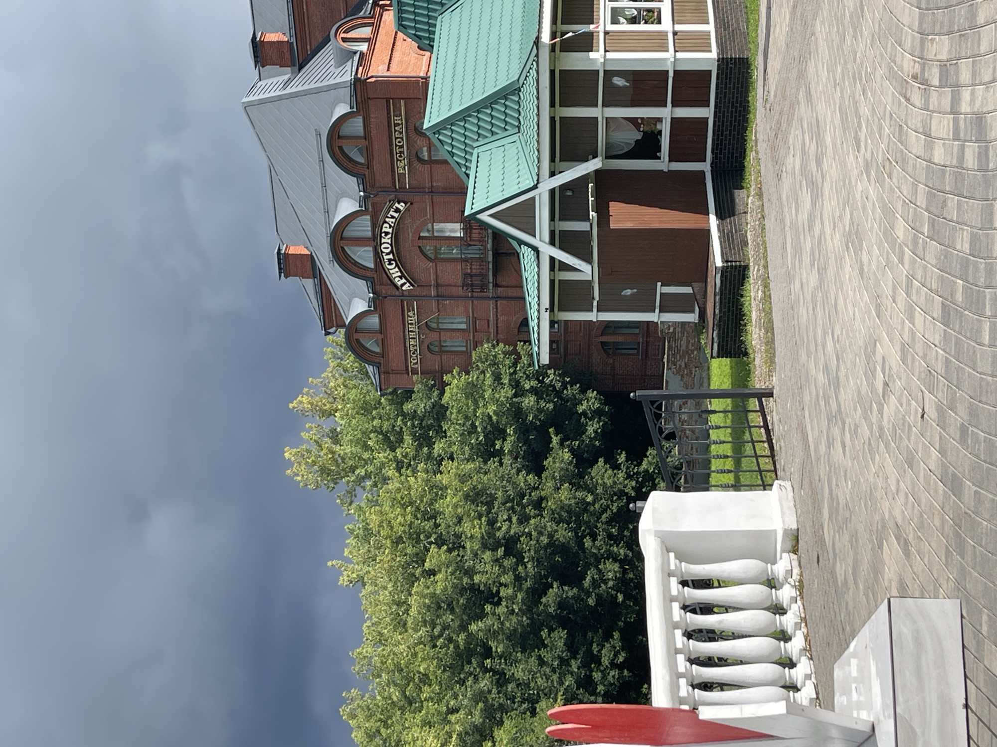 The image depicts the exterior of a building complex in Sergiev Posad, Russia, identified by signage as "ГОСТИНИЦА АРИСТОКРАТЬ РЕСТОРАН" (Hotel Aristocrat Restaurant). The main building is constructed of red brick with a grey, mansard-style roof featuring arched dormer windows. A smaller, detached structure with a green metal roof is situated in front of the main building. The foreground shows a paved walkway, a white balustrade, and a black metal gate. Lush green trees form a backdrop on the left side of the image, contrasting with a dark, overcast sky. No people are visible in the scene. The setting appears to be a sunny day despite the dark clouds, with shadows cast by the buildings and trees.