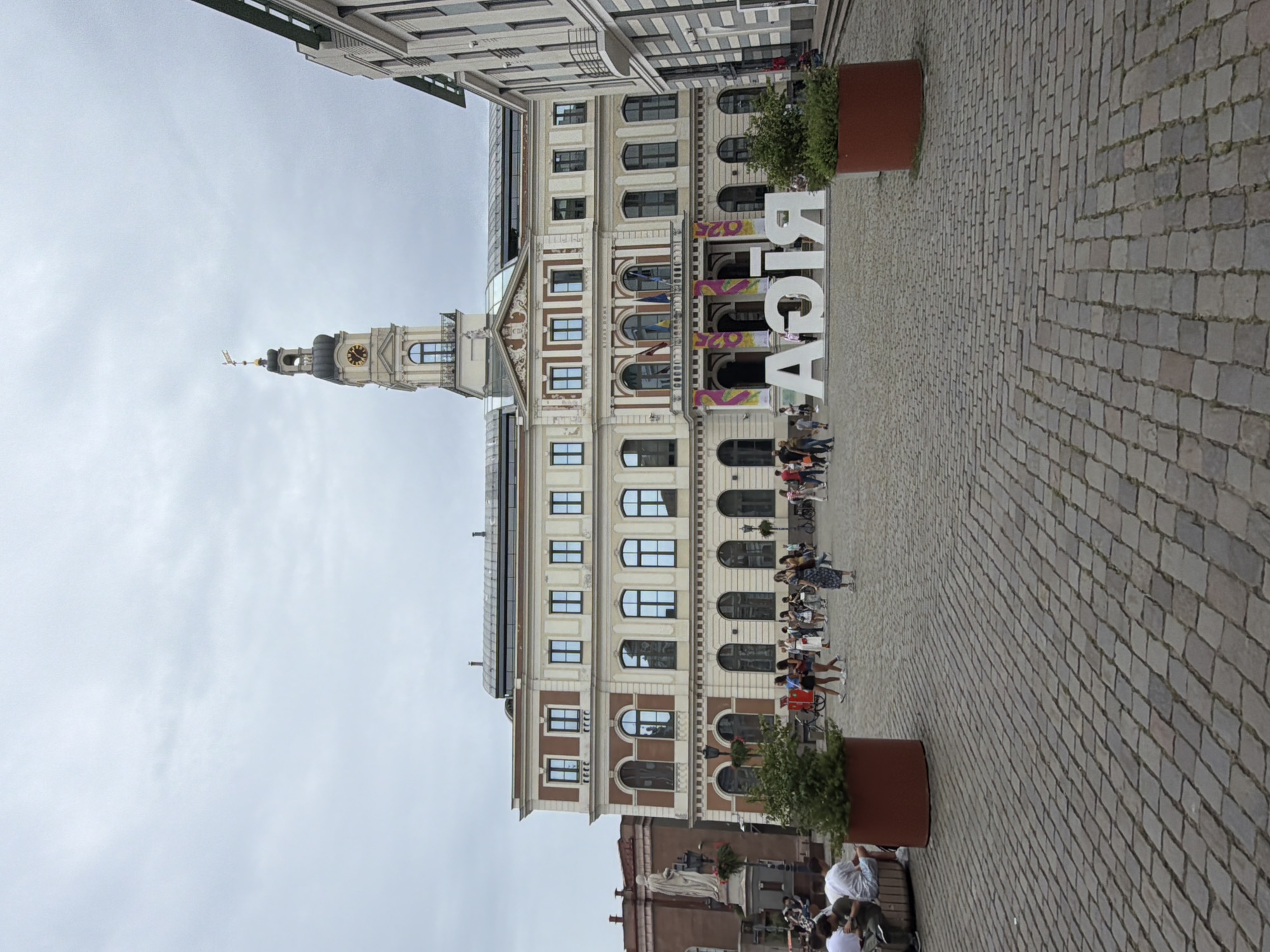 The image shows the Riga City Council building in Riga, Latvia. The building is a large, ornate structure with a prominent clock tower. In front of the building, there is a large, white "RIGA" sign. Several people are walking around the square, some sitting on benches. The ground is paved with cobblestones. The sky is overcast.