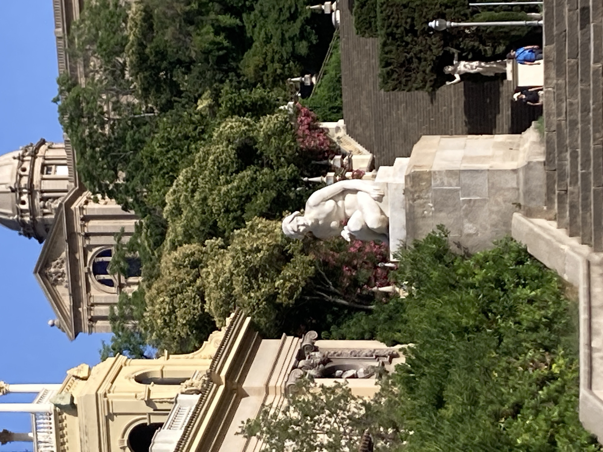 The image captures a view of the Palau Nacional on Montjuïc hill in Barcelona, Spain. In the foreground, a tiered stone staircase ascends towards the building, flanked by lush green hedges and trees. A white marble statue of a seated female figure is prominently placed on a pedestal near the bottom of the staircase. Further up the stairs, another statue of a standing female figure is visible, and two individuals, a woman and a man, are walking up the steps. The Palau Nacional itself, a grand neoclassical building with a large dome, forms the backdrop, partially obscured by mature trees. The sky above is a clear, bright blue.