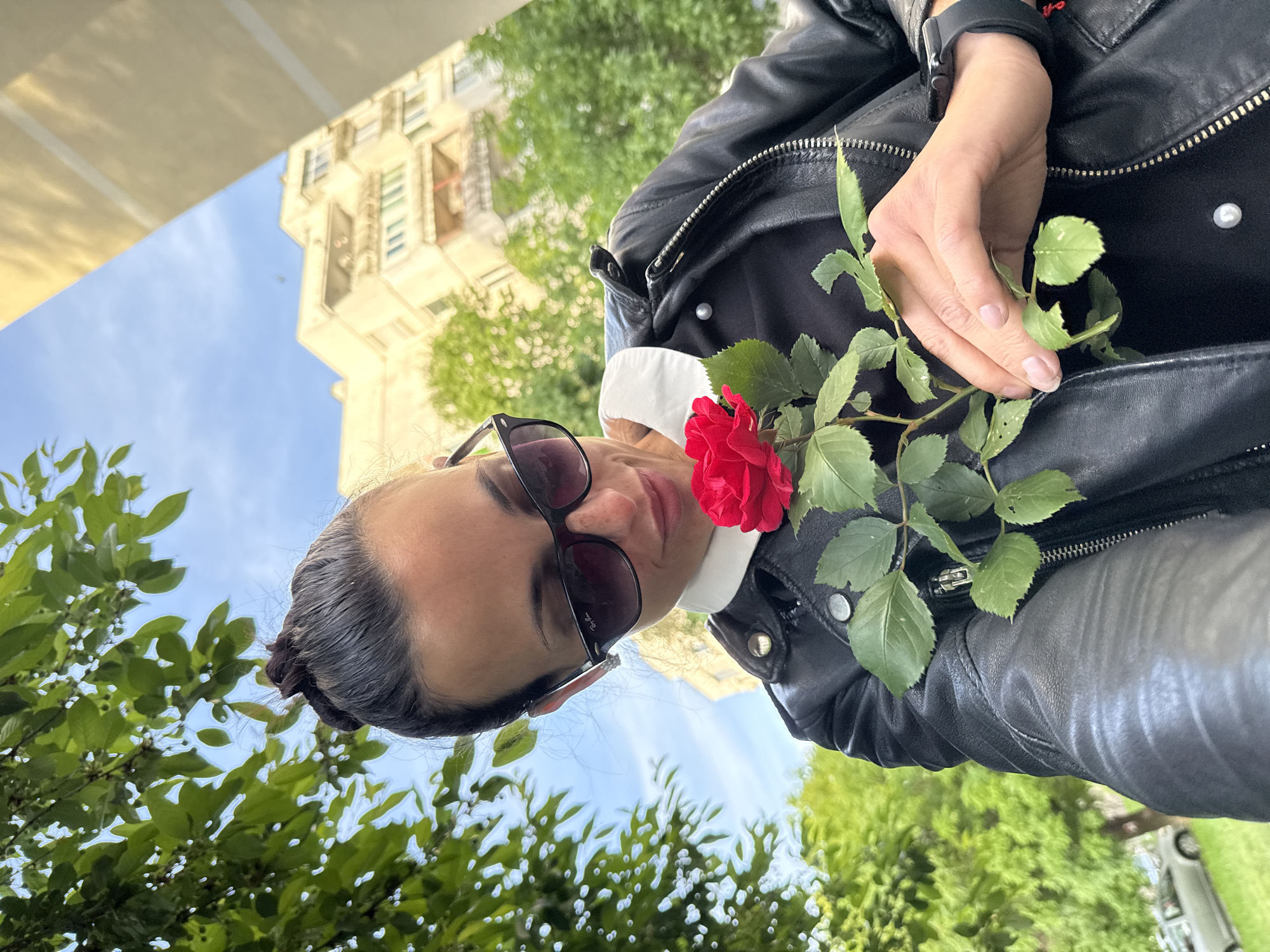 A woman with dark hair pulled back into a bun is holding a red rose with green leaves. She is wearing sunglasses and a black leather jacket over a white collared shirt. The background includes green trees and a building. The location is not explicitly stated as Pleven, Bulgaria, based on the provided image alone.
