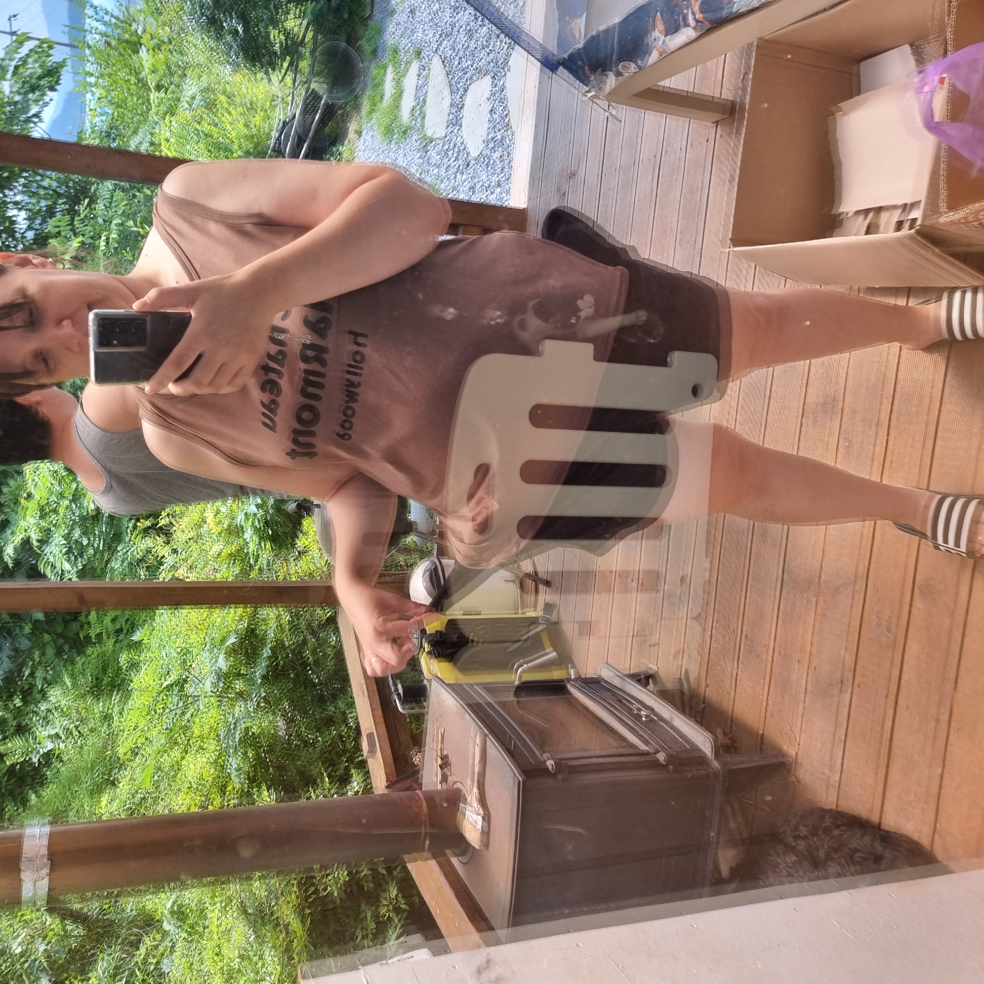 In Dunnae-myeon, South Korea, on a wooden deck, a woman in a brown tank top and dark shorts is taking a selfie. She is holding a smartphone in her right hand. To her left, a dark wood-burning stove with a metal chimney stands on the deck. There is a brush on top of the stove. Behind the woman, another person in a grey tank top is visible. The deck is surrounded by lush green trees and foliage. A gravel path with stepping stones leads away from the deck. A cardboard box filled with papers is visible in the foreground. The scene appears to be an outdoor living space.