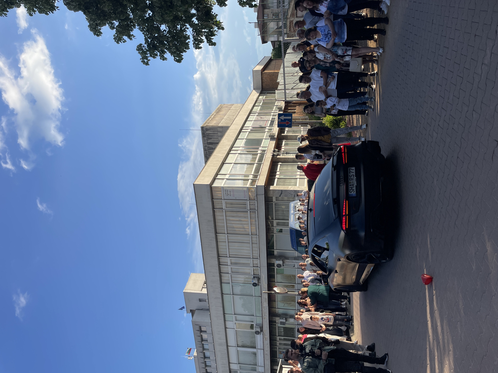 The image shows a scene in Zlatograd, Bulgaria. A large group of people is gathered on a brick-paved street in front of a multi-story building. The building has a concrete facade with rows of windows. A black car is parked on the street, partially blocking the view of some of the people. There is also a red balloon lying on the pavement near the car. In the foreground, there are uniformed personnel. Above the building, the sky is blue with scattered clouds. Tree branches are visible in the upper left corner of the image.