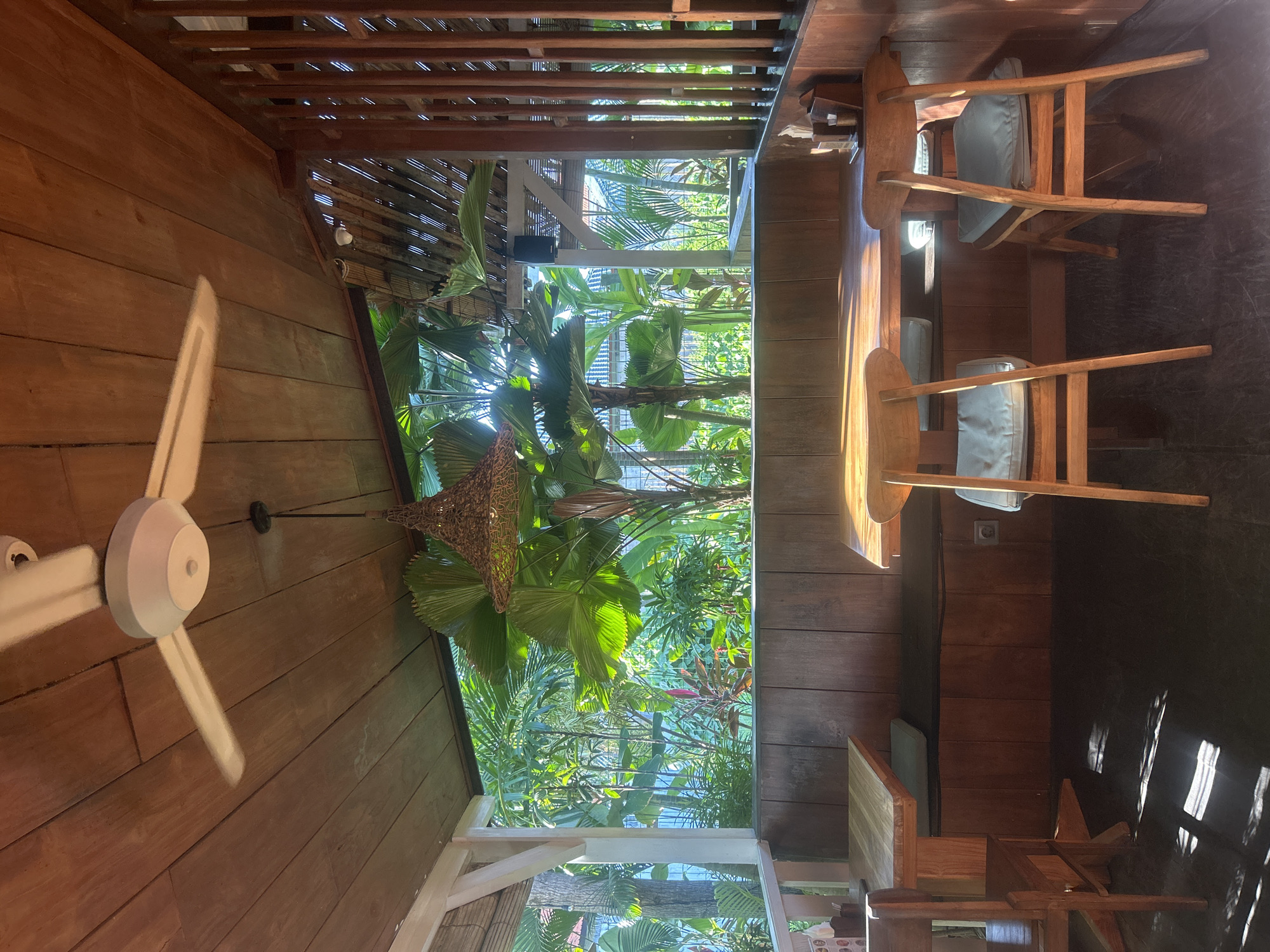 This is a photograph taken in North Kuta, Indonesia, of an outdoor seating area of a restaurant or cafe. The scene is characterized by its use of natural materials, predominantly wood, and lush tropical greenery visible through an open-air partition.
In the foreground, two wooden tables are set up. The table on the left is partially visible, with a wooden chair beside it. The table on the right is more prominently featured, with two matching wooden chairs positioned around it. Each chair has a light blue cushion. The tables are set for dining, with a napkin holder and possibly cutlery visible on the right table.
The seating area is covered by a wooden ceiling with visible beams and planks. A white ceiling fan is mounted on the wooden ceiling, its blades creating a blur due to motion. A woven lampshade hangs from the ceiling between the fan and the view outside.
Behind the tables, a wooden bench seat is installed along a wooden wall. Above the bench, a large opening reveals a vibrant view of a tropical garden. Large, fan-shaped leaves of palm trees are dominant, interspersed with other green foliage and some smaller plants. The sunlight filters through the leaves, creating dappled patterns on the ground and the wooden surfaces.
The walls are constructed of dark wood planks. On the right side of the image, a slatted wooden partition adds to the open, airy feel of the space. The floor is a dark, flat surface, likely concrete or stone, with dramatic light patterns cast by the sun through the overhead structure and foliage. The overall atmosphere is relaxed and tropical, suggesting a pleasant dining or lounging experience in a warm climate.