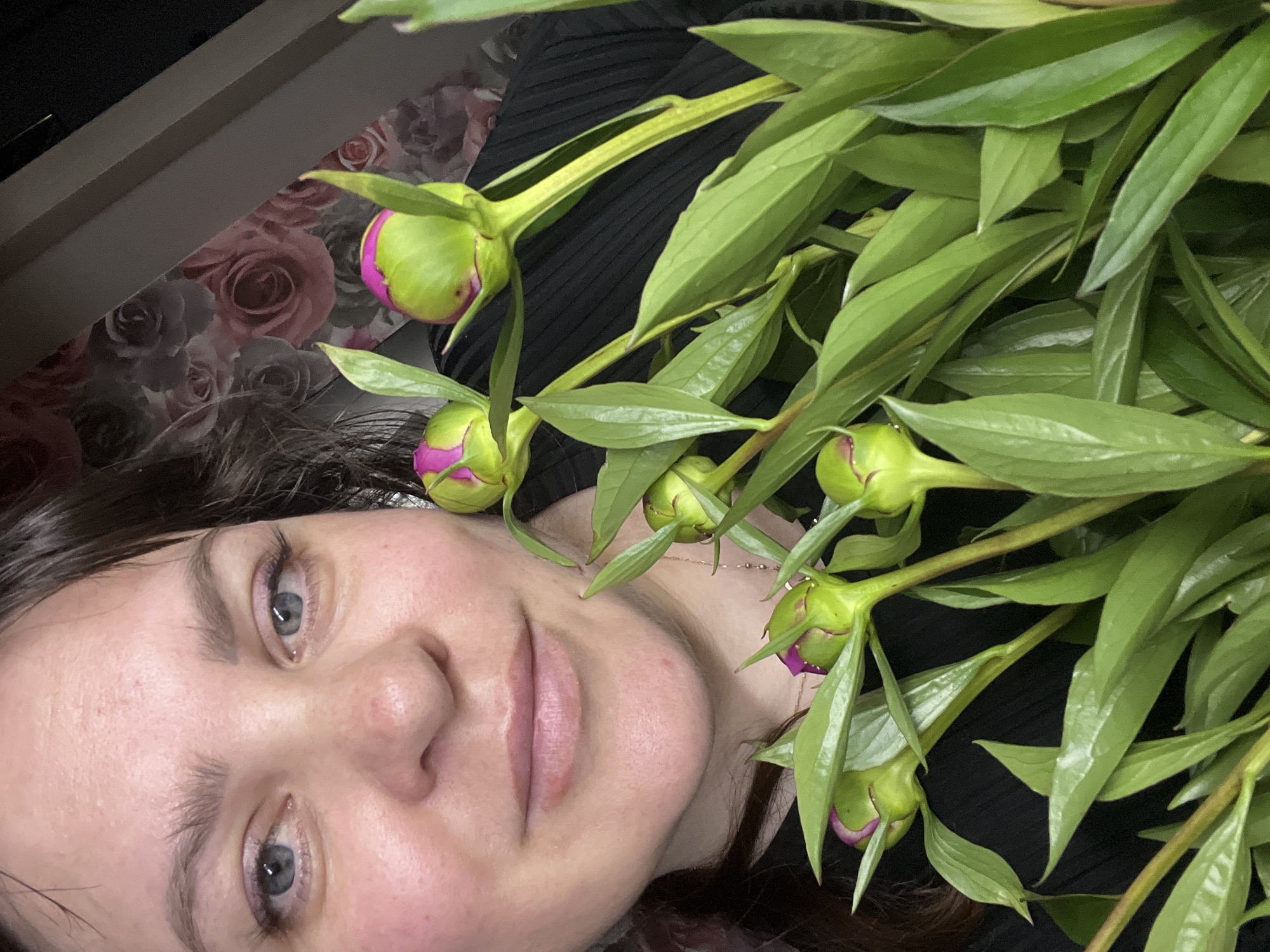 A woman with dark hair and blue eyes is holding a bouquet of peonies in front of her face. She is wearing a black top. The background is a floral wallpaper with pink and grey roses, and a diagonal brown beam. The image appears to be taken indoors, likely in a residential setting, in Noviy, Russia.