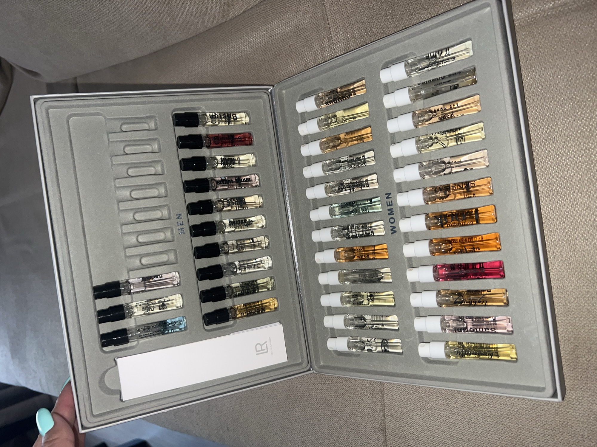 The image shows a close-up overhead view of an open beauty product set, likely a perfume sample collection, placed on a textured beige surface, possibly a couch or carpet. The set is housed in a gray, segmented case, with rows of small glass vials filled with various colored liquids. The case is divided into sections labeled "MEN" and "WOMEN".
In the "MEN" section, which is in the upper part of the image, there are two rows of vials. The top row contains seven vials, three of which are partially visible on the left. The second row contains ten vials, six of which are fully visible and four are partially visible on the right.
The "WOMEN" section, in the lower part of the image, also has multiple rows of vials. The first visible row has ten vials, all of which are mostly or fully visible. The second visible row has seven vials, all of which are mostly or fully visible.
On the left side of the "MEN" section, there is a white card with a black logo that appears to be "LR HEALTH & BEAUTY". A person's hand with light blue nail polish is visible in the upper left corner, reaching towards the set. The background is out of focus and appears to be a domestic setting.
There is no information in the image that indicates a location in Plovdiv, Bulgaria.