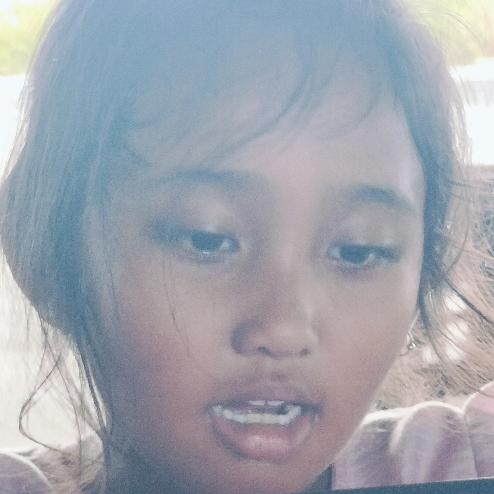 A close-up, brightly lit portrait captures a young person, likely a girl, with dark hair, a round face, and dark eyes, looking downwards. Her mouth is slightly open, revealing small, spaced lower teeth. The image is significantly overexposed, particularly in the background, which is blurred into abstract light blue, white, and faint yellow-green hues, suggesting strong natural daylight or an outdoor setting.
The subject's skin tone appears to be of Asian descent, consistent with the location context of Keuniree, Indonesia. Her dark hair falls around her face. A small earring is visible on her right ear (viewer's left). The image has a soft, hazy quality, making specific background details indistinguishable. No specific activity is discernible other than being the subject of a photograph, and her expression is neutral or contemplative as she gazes downward. No text is visible in the image. The overall impression is a soft, sun-drenched portrait of a child.