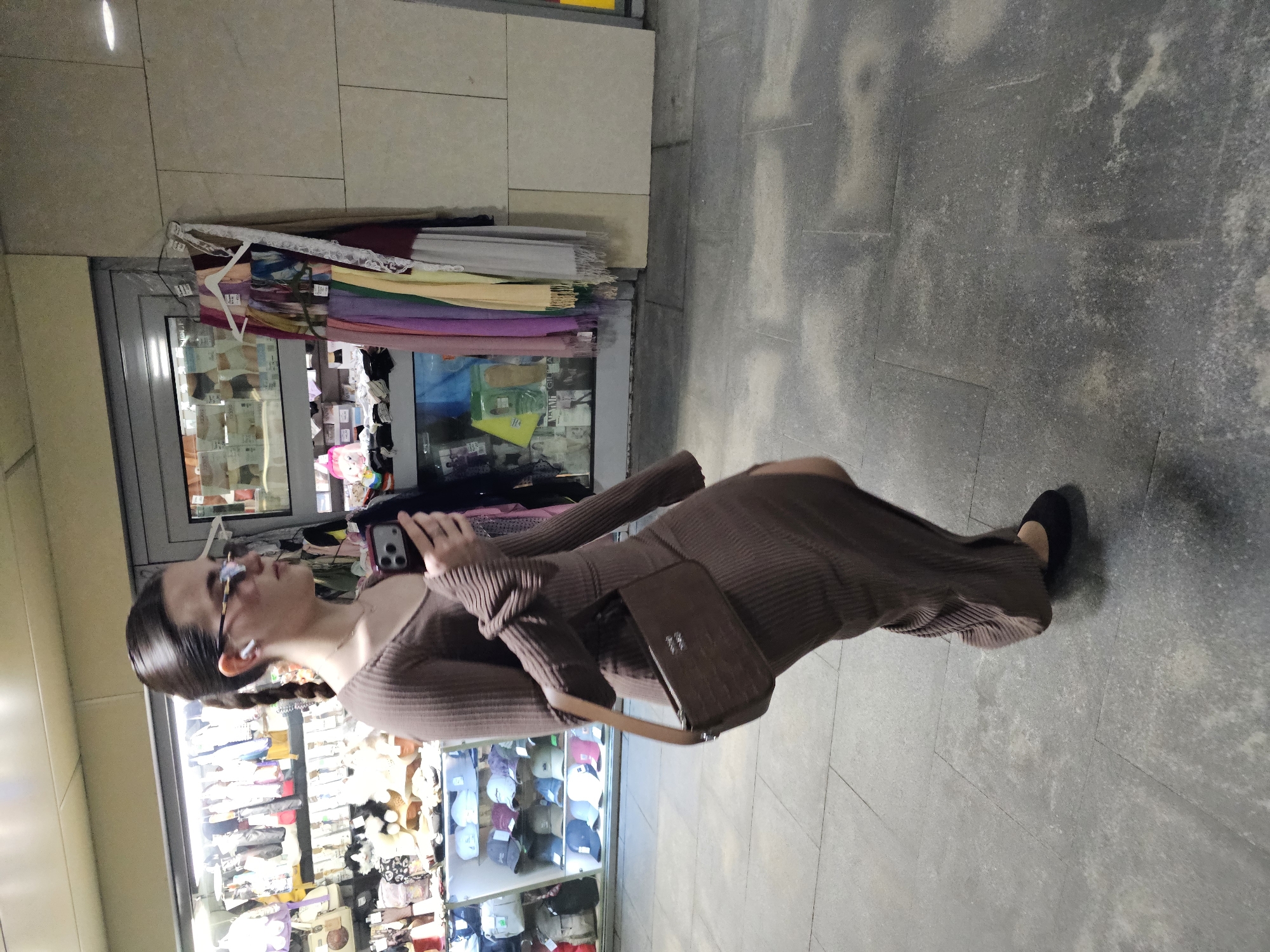 A woman with braided hair, wearing a brown long-sleeved dress and black shoes, is standing and holding a smartphone. She is near the front of a storefront with merchandise displayed inside, including hats and plush toys. There are garments hanging on display near the top of the storefront window. The location appears to be a retail area, with a concrete floor. The city and specific region are not identifiable from the given image.