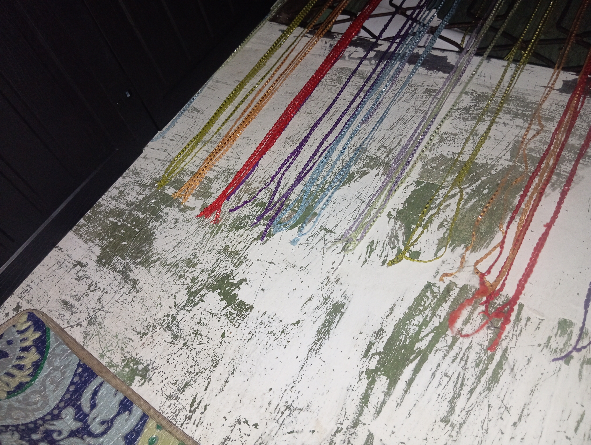 The image depicts a collection of colorful, thin strands, possibly strings or threads, laid out on a weathered, white surface with patches of green paint showing through. The strands are arranged in parallel lines and vary in color, including yellow, orange, red, purple, and blue. Some strands appear to be made of individual beads or have a textured appearance, while others are smooth and braided. A dark, wooden surface is visible in the upper left corner, and a patterned fabric or mat is partially visible in the lower left corner. The overall impression is of craft materials or decorative elements arranged for display or use. The image does not contain any information about the specific location in Rejosari, Indonesia, or any individuals present.