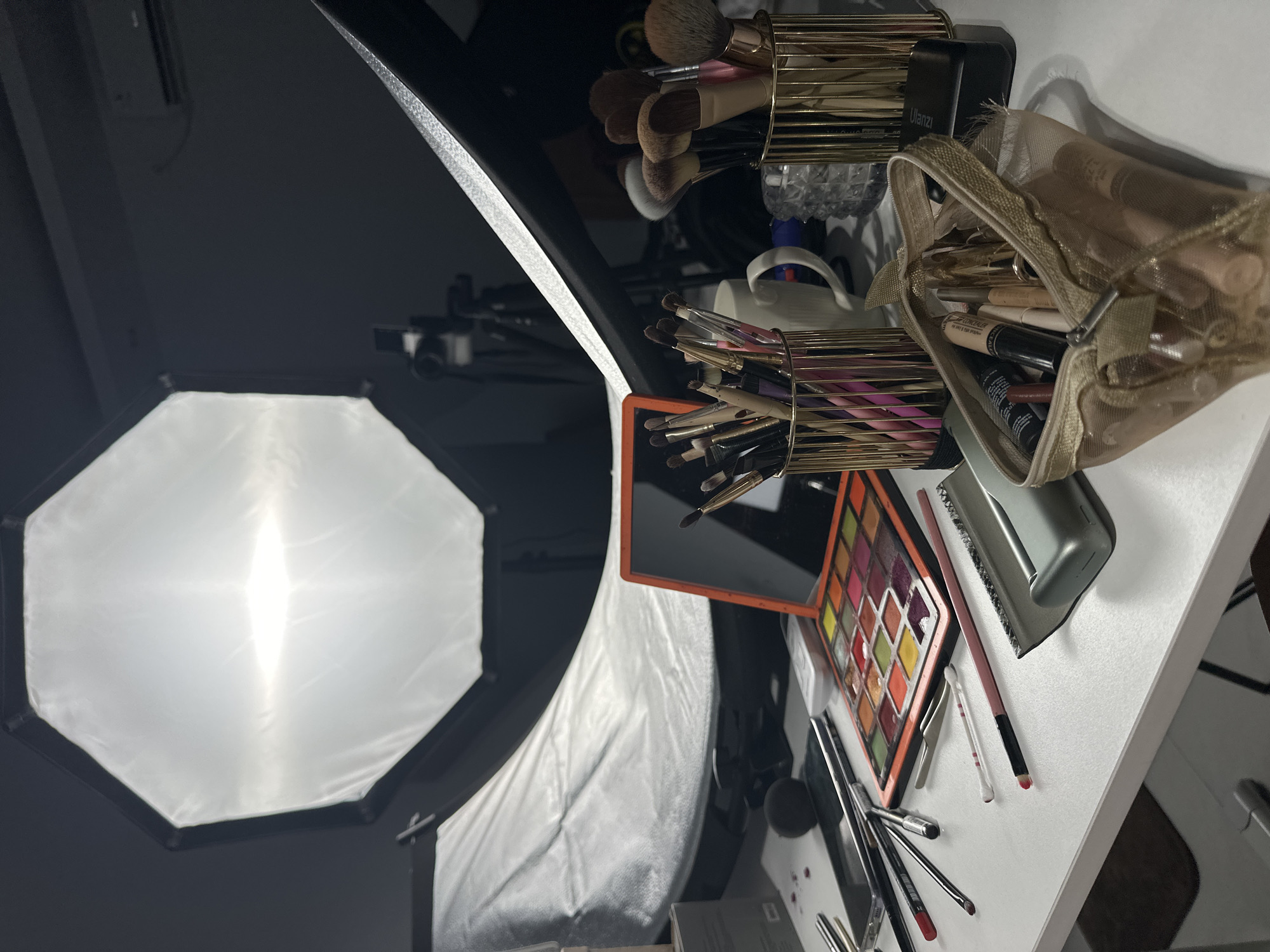 The image depicts a cluttered workspace. A white softbox, used for lighting, is positioned on the left. On a white surface, likely a table, there are various makeup items: multiple makeup brushes in gold containers, a makeup bag filled with cosmetics, a makeup palette with various colors, individual makeup brushes, cotton swabs, and tweezers. The location is North Kuta, Indonesia. There are no people in the image.