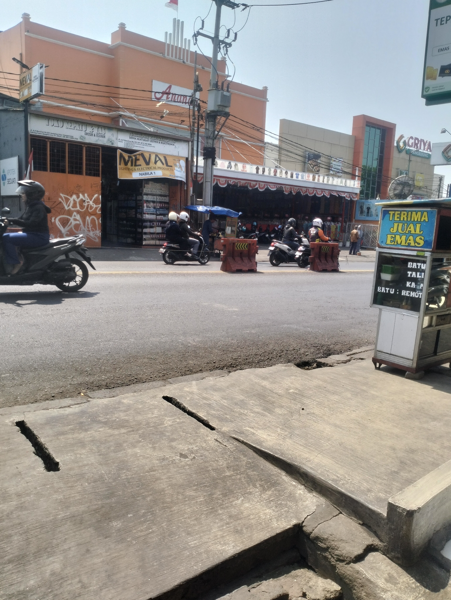 The image depicts a street scene in Kel. Pasir Jati, Indonesia, under a clear sky. In the foreground, a concrete sidewalk with two linear indentations is visible, bordering a paved road. Several motorcycles are on the road. One motorcycle, ridden by a person wearing a dark jacket and helmet, is moving away from the viewer on the left side of the image. Further down the road, two motorcycles, each carrying two individuals wearing helmets, are also traveling. On the right side of the road, a mobile kiosk with a blue and yellow sign reading "TERIMA JUAL EMAS" (accepts gold sales) is parked. The kiosk has text on its side indicating "BATU TALI KA P BATU : REMOT".
Behind the road, a row of buildings lines the street. The building on the left is an orange structure with a sign that reads "MEVAL LIGHTING & ELECTRICAL PRODUCTS". Next to it, there is a store with open shutters displaying shelves of merchandise. The building to the right is a modern multi-story structure with a prominent sign that includes "GRIYA" and appears to be a retail establishment. Power lines and utility poles are visible crisscrossing above the street and buildings. The overall environment suggests a commercial area on a sunny day.