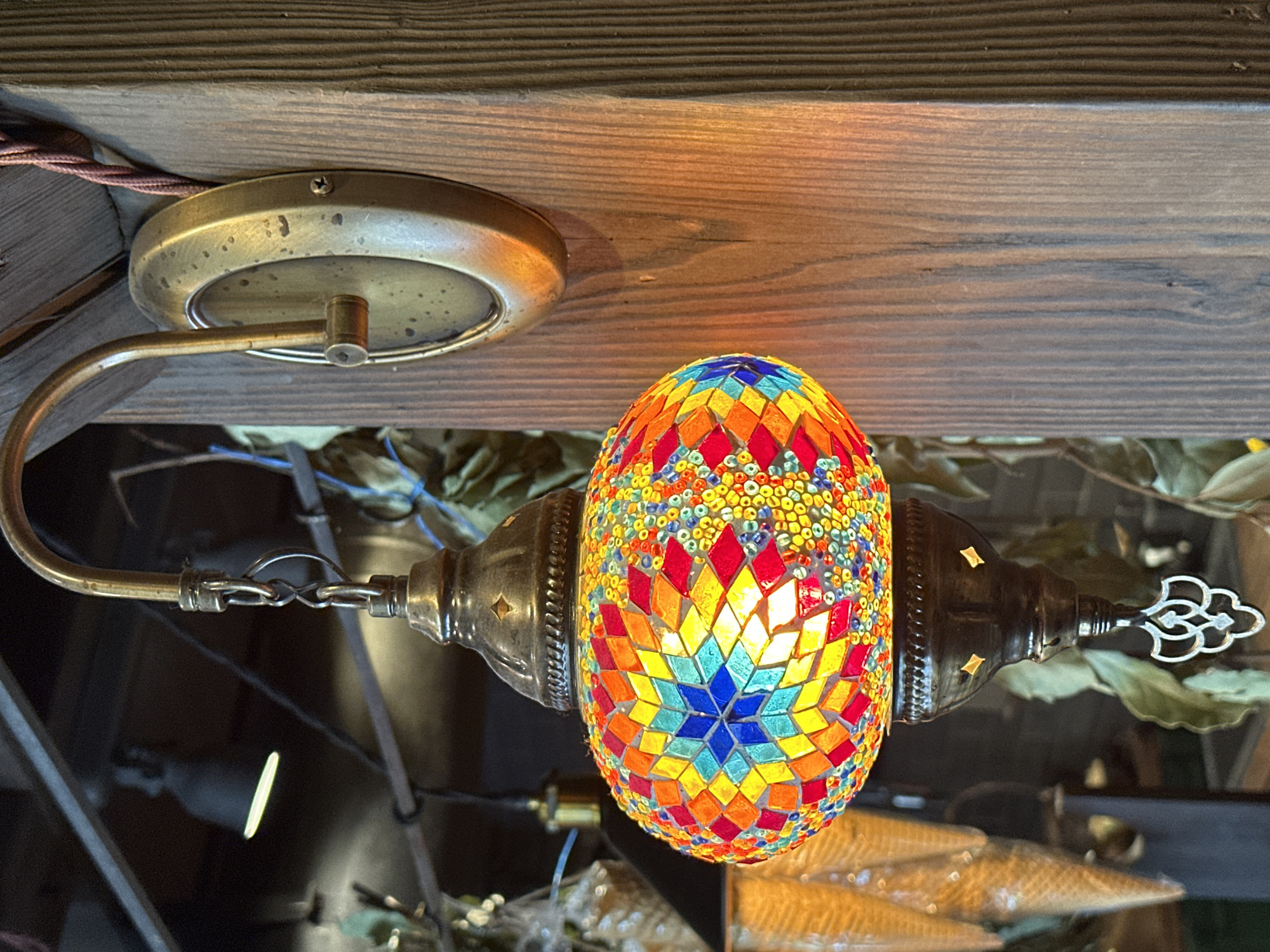 A detailed view captures a hanging lamp fixture in what appears to be an interior setting, possibly a shop or home in Новоглаголево, Russia. The main subject is an ornate, mosaic-covered lamp with an egg-shaped shade. It features a geometric pattern made from colorful glass or tile pieces, primarily red, orange, yellow, blue, and green. The light from within illuminates the design.
The lamp is suspended from a curved, brass-colored metal arm connected to a circular ceiling mount that is also made of brass. There is a visible screw in the ceiling mount. The lamp is attached to the arm via a chain and ornate metal fittings at either end of the bulb. The background and surroundings are somewhat obscured, with glimpses of other objects and greenery suggesting a decorated space. The lamp is positioned against a dark, possibly wooden surface.