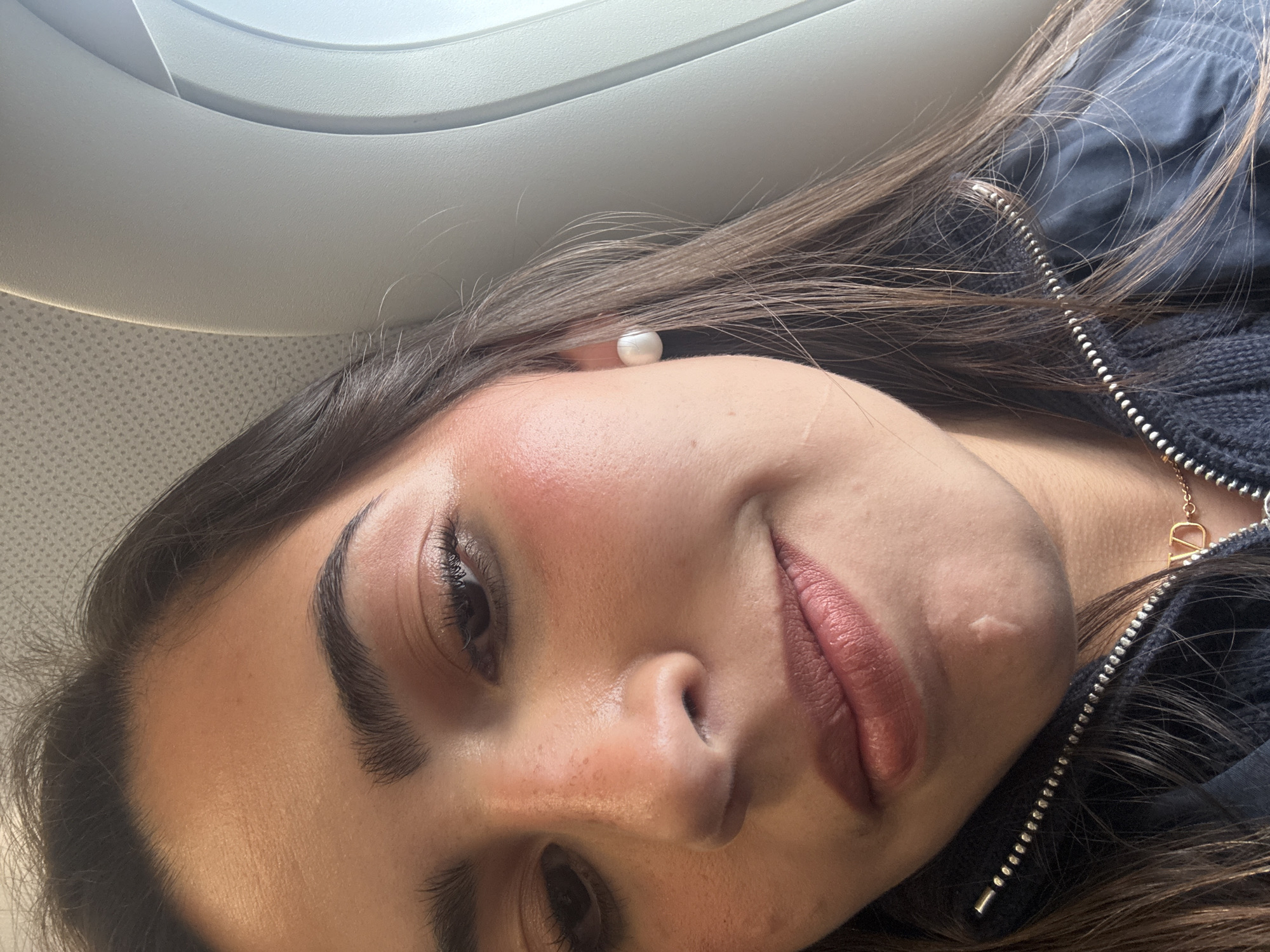 A close-up, tilted shot shows a young woman's face. She has brown hair, dark eyebrows, and fair skin. She is wearing a pearl earring. Her lips are a rosy color. A scar is visible on her chin. She's wearing a black jacket with a silver zipper and a gold necklace with a pendant shaped like a capital "D". Part of an airplane window is visible in the upper left corner of the frame. It is impossible to determine the specific location of the image in San Antonio based on the given information.