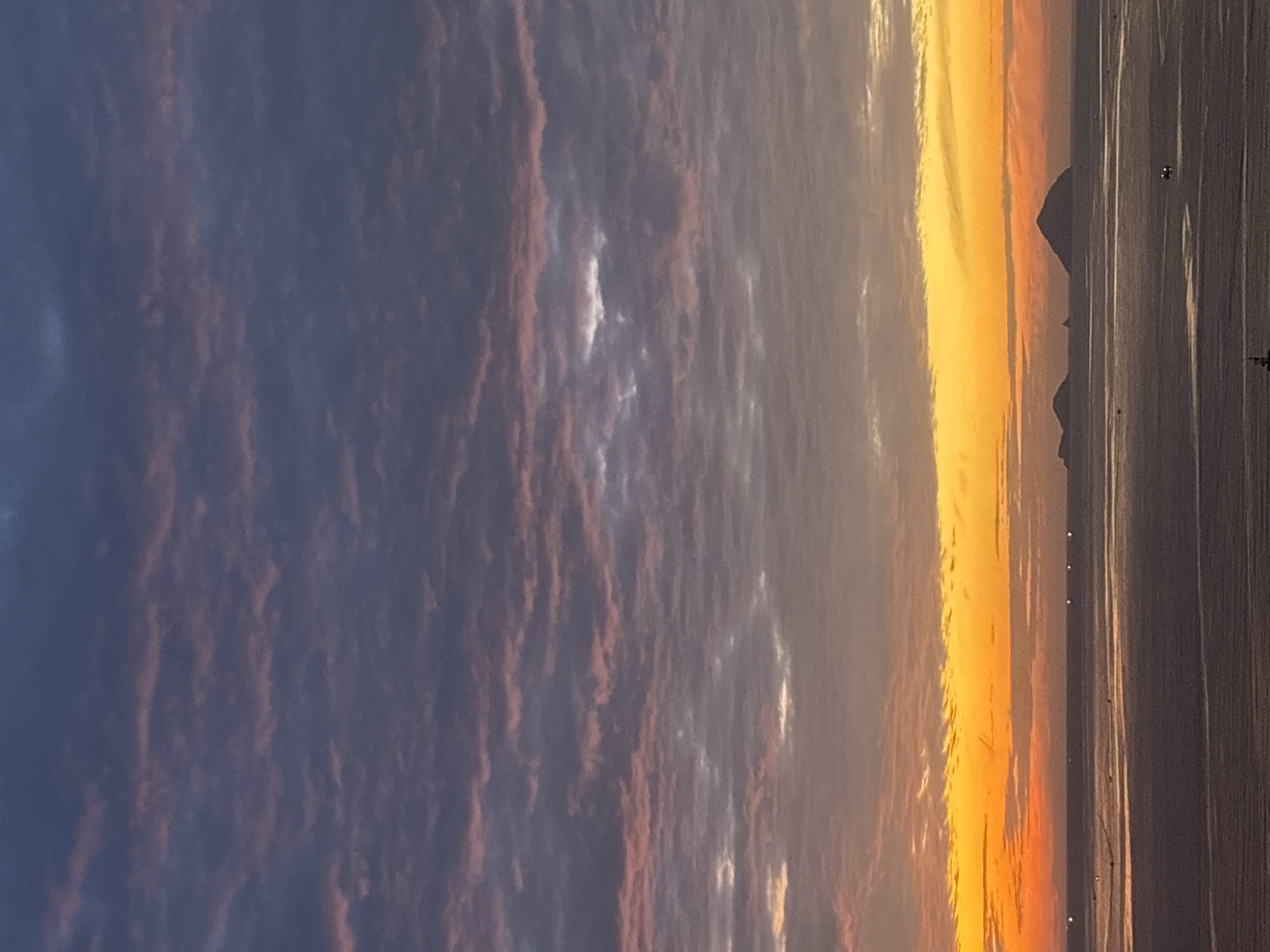 The image depicts a twilight scene over the ocean near Nha Trang, Vietnam. The sky is filled with striated clouds, transitioning from deep indigo and grey at the top to vibrant orange and yellow near the horizon. Several islands, silhouetted against the brightly lit sky, are visible in the distance. The ocean is calm, reflecting the colors of the sunset. Small lights, likely from boats, are scattered across the water. The overall impression is one of serene natural beauty at the end of the day.