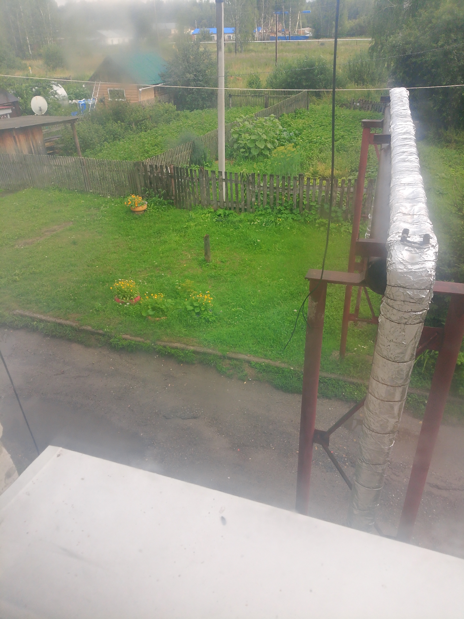 The image shows an outdoor scene in Asino, Russia, likely a residential area. In the foreground, a white surface, possibly a windowsill or railing, partially obscures the view. A large, insulated pipe, wrapped in reflective foil, is prominently featured on the right side of the image, supported by a red metal structure. Behind the pipe, a wooden fence encloses a grassy yard with some potted flowers. Beyond the fence, a small wooden house with a green roof and a satellite dish is visible, surrounded by vegetation. Further in the background, trees and distant buildings suggest a rural or semi-urban environment. The overall impression is one of a slightly overcast day, with evidence of rain or mist contributing to a hazy atmosphere. There are no people visible in the image.