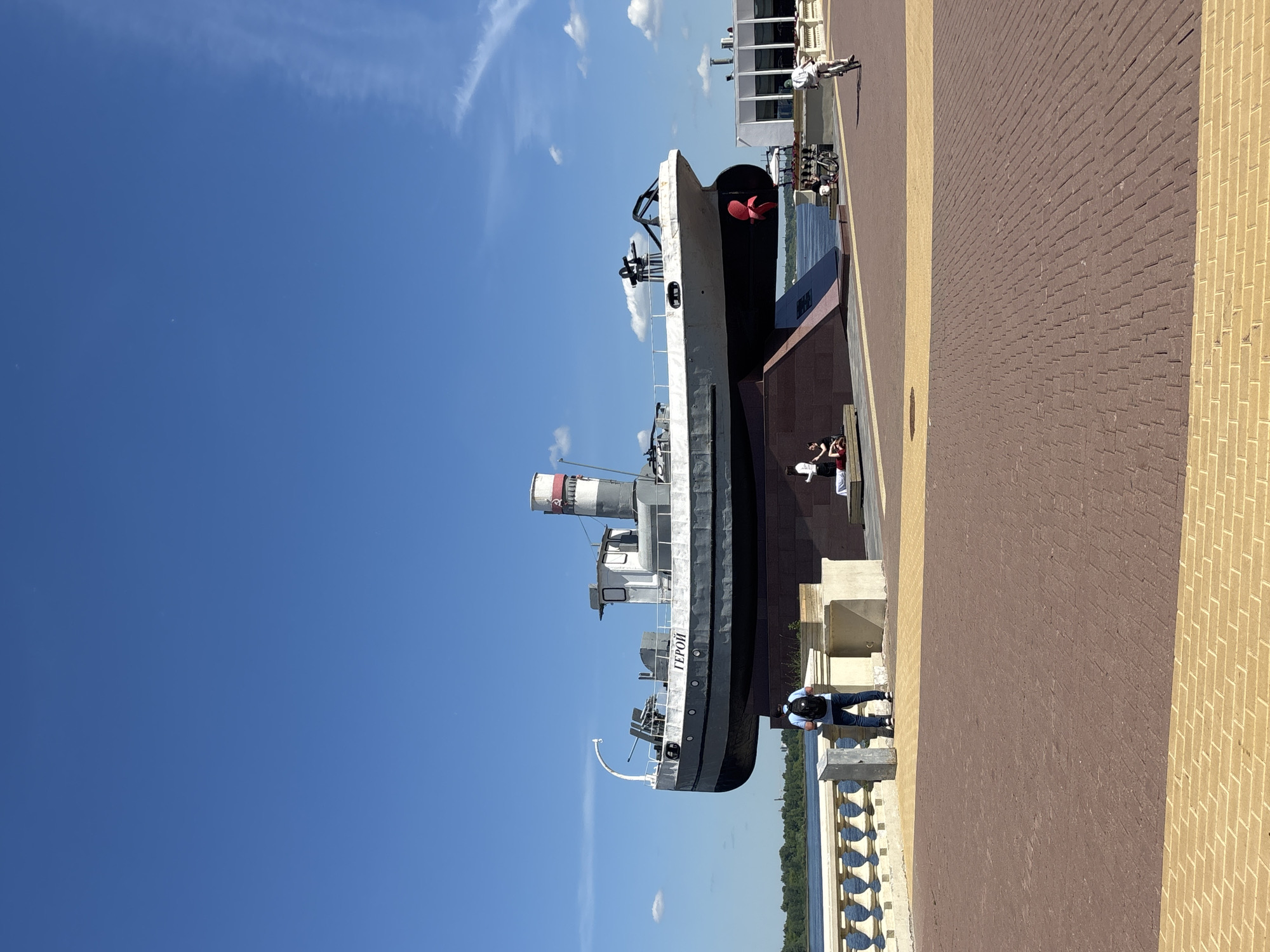 The image displays a sunny daytime scene along a waterfront, featuring a large ship docked alongside a paved promenade. The central subject is a grey and black ship, which appears to be a historical vessel or a museum exhibit. Its stern, visible on the right, features a prominent red propeller. The ship has a white superstructure, a tall smokestack with red and white stripes, and railings along its decks. The word "ГЕРОЙ" (HERO) is clearly visible in white letters on its hull.
In the foreground, a wide promenade is paved with reddish-brown and yellow brick-like pavers. A white decorative railing separates the promenade from a body of water, visible in the background, lined with distant trees and faint structures. Several people are present: one individual pushes a scooter or trolley near the right edge, two people are seated on a bench or structure adjacent to the ship's stern, and another person wearing a backpack stands near the white railing overlooking the water. The sky is a clear, bright blue with a few scattered white clouds, indicating pleasant and sunny weather. The overall atmosphere suggests a relaxed, leisurely setting.