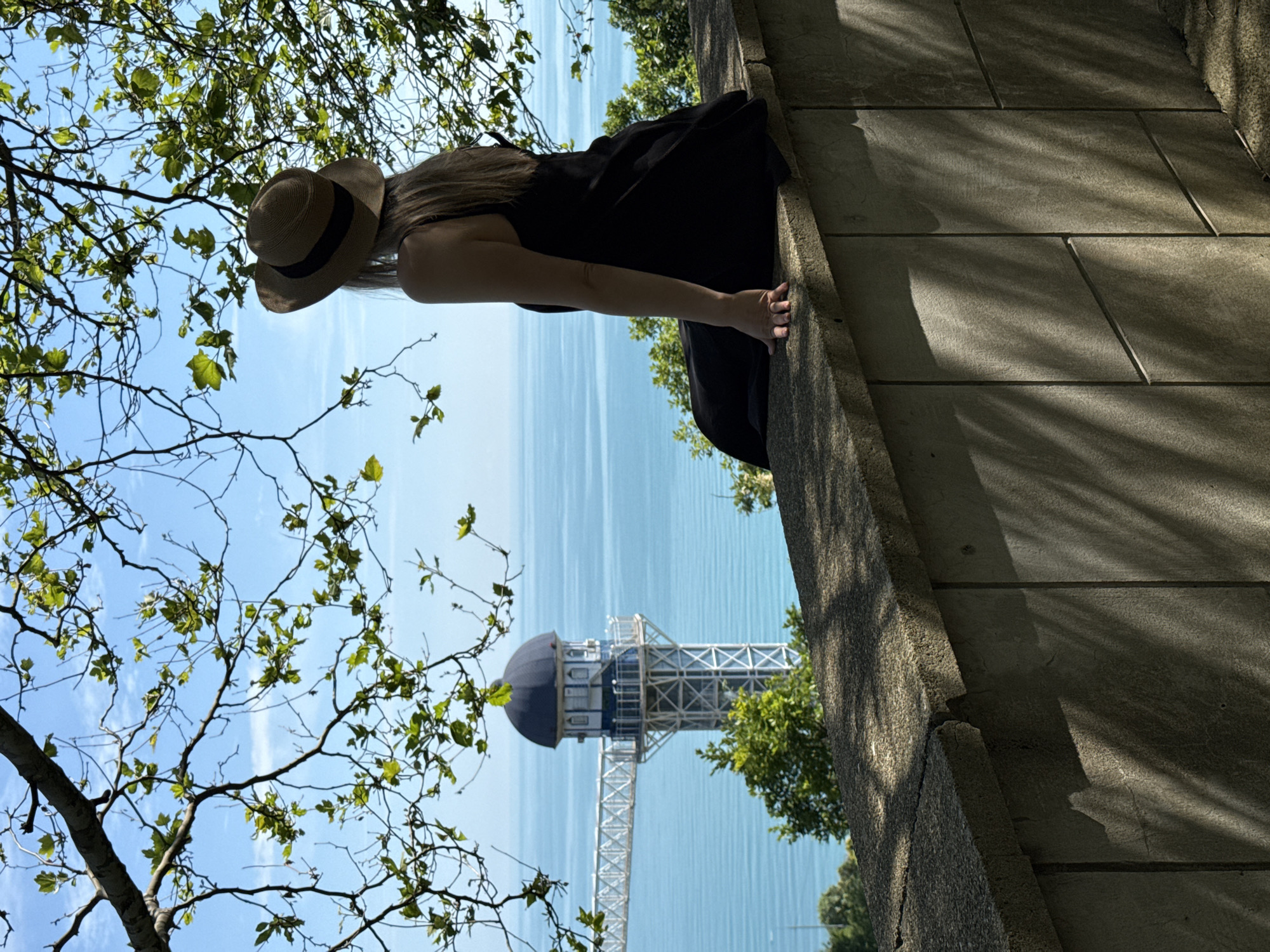 A person wearing a straw hat and a black dress sits on a stone wall overlooking the sea in Nebug, Russia. The person is facing away from the camera, looking towards a tall, white tower with a blue dome that extends out over the water on a metal bridge. Green trees and branches frame the top and sides of the image, with dappled sunlight casting shadows on the wall and ground. The sea is a vibrant turquoise, with gentle waves visible. The sky is clear and blue with a few wispy clouds.
