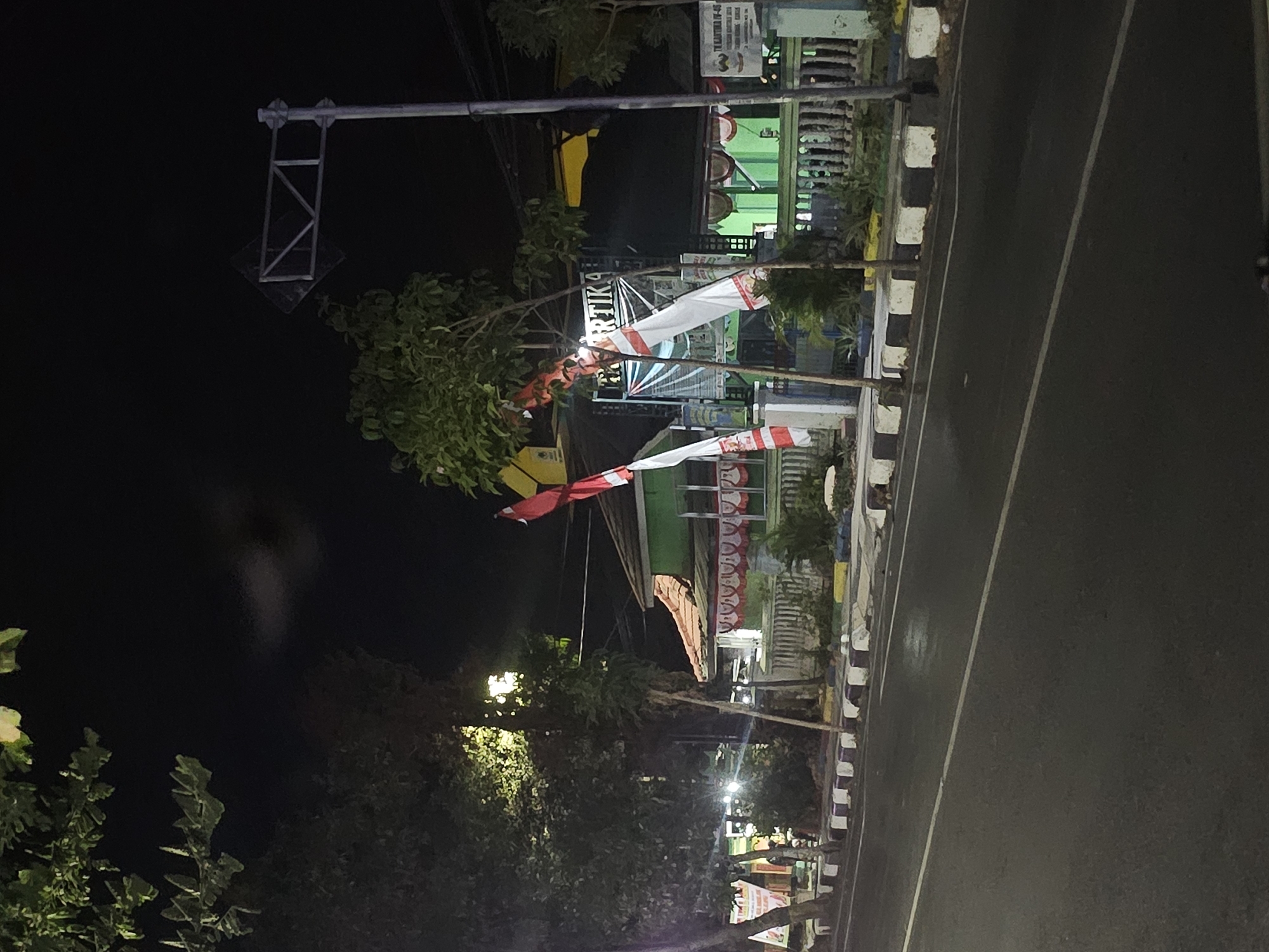 The image, taken at night, shows a street scene in Lamongan, Indonesia. In the foreground, a dark asphalt road with a white dividing line runs across the bottom of the frame. To the left, a sidewalk with a border of alternating white and purple blocks is visible, lined with trees and some low vegetation. Behind the sidewalk, a green building with a tiled roof and a white fence is illuminated, with decorative red and white banners hanging from the roofline and flags on poles. The building appears to be a commercial establishment, with the word "PARTIKA" visible in large letters above the entrance, along with other text on signs. The overall atmosphere is that of a quiet street at night, possibly decorated for a local celebration given the flags and banners.