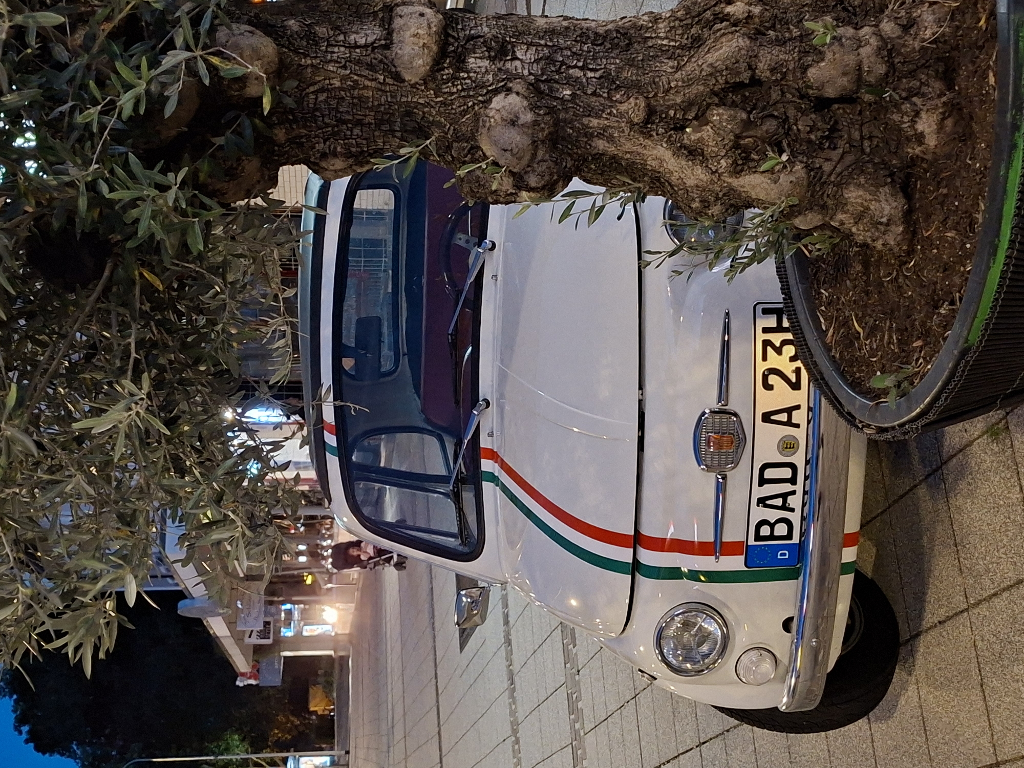 The image shows the front of a white Fiat 500 decorated with Italian flag racing stripes, parked on a paved street at night. The car's license plate reads "BAD A 23H". A large, gnarled olive tree stands to the right of the car, partially obscuring it. In the background, there are streetlights, buildings, and a person pushing a stroller with a child. The setting appears to be a pedestrian zone in a European city, consistent with Baden-Baden, Germany, given the license plate prefix "BAD".