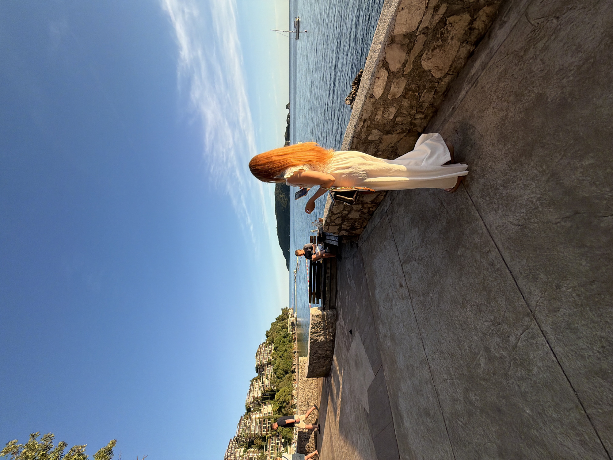 This image depicts a vibrant coastal scene in Budva, Montenegro, captured during the late afternoon. The warm, golden sunlight illuminates the setting, casting soft shadows and highlighting the deep blue sky, which features a few wispy white clouds.
The primary subject is a person with long, flowing reddish-orange hair, dressed in a light, possibly white or cream-colored, long dress. They stand on a paved promenade, facing the expansive blue sea, holding a mobile phone in their hands, perhaps taking a photograph or viewing content. A small bag hangs from their shoulder.
The promenade is bordered by a robust stone wall that separates it from the calm waters of the Adriatic Sea. In the middle distance, a white sailboat with a tall mast is visible on the water. Across the bay, a lush green hillside rises, densely covered with trees and dotted with numerous multi-story buildings, typical of terraced Mediterranean architecture, extending up the slopes.
Further along the promenade, towards the left, other individuals are present. Two people are seated on a dark wooden bench, appearing to relax and enjoy the view, while another man in a dark t-shirt and light shorts walks by. The overall atmosphere is peaceful and leisurely, suggesting a pleasant day spent by the sea. No visible text is present in the image.