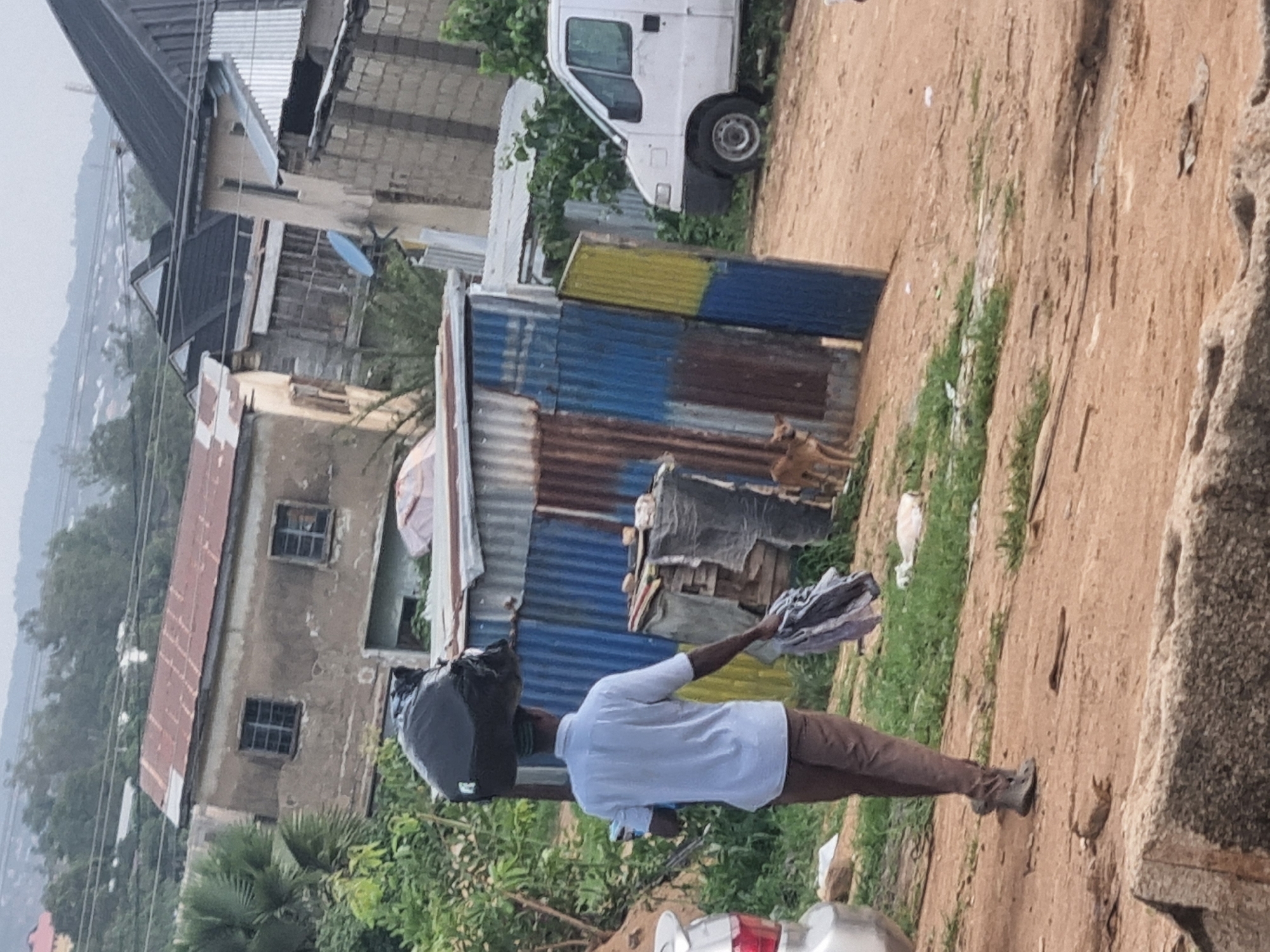 A man is walking away from the camera in an outdoor scene, carrying a large black garbage bag on his head. He is wearing a light blue short-sleeved shirt and brown pants. He is also holding a bundle of folded grey and white fabric in his right hand. The man is walking on a dirt path, with green grass and some scattered debris to his left. Behind him, there is a corrugated metal structure with blue and yellow panels. To the right of the metal structure, a small brown dog is standing and looking towards the camera. Further to the right, a white van is parked. In the background, there are several buildings with various roofing materials, some appear to be in disrepair. The overall environment suggests an urban or semi-urban setting in Jos, Nigeria, possibly a less developed area. The sky is overcast.