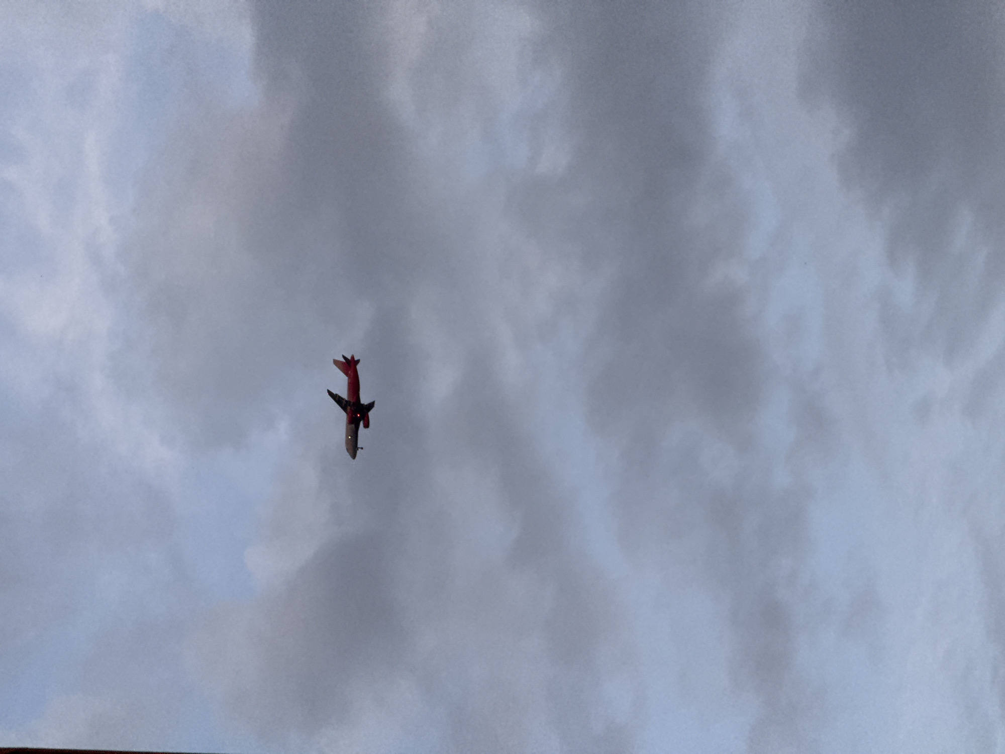 The image captures a small, red and grey model airplane flying upside down against an overcast sky. The airplane, likely a radio-controlled (RC) aircraft, is depicted in an inverted flight maneuver, with its red top surface and tail fins pointed downwards and its grey underside facing upwards. The wings are visible, contributing to the distinct silhouette of a fixed-wing aircraft.
The scene is set outdoors under a cloudy sky, characterized by varying shades of grey and light blue, suggesting a heavily overcast or stormy day. There are no clear indications of the time of day, but the ambient light suggests daytime. No specific landmarks or geographical features from Saint Petersburg, Russia, are visible within the frame, apart from a sliver of a dark, possibly building-related, structure at the very bottom edge of the main image. The activity is the flight of the model airplane, showcasing an aerobatic maneuver. There is no visible text or any human figures in the image.