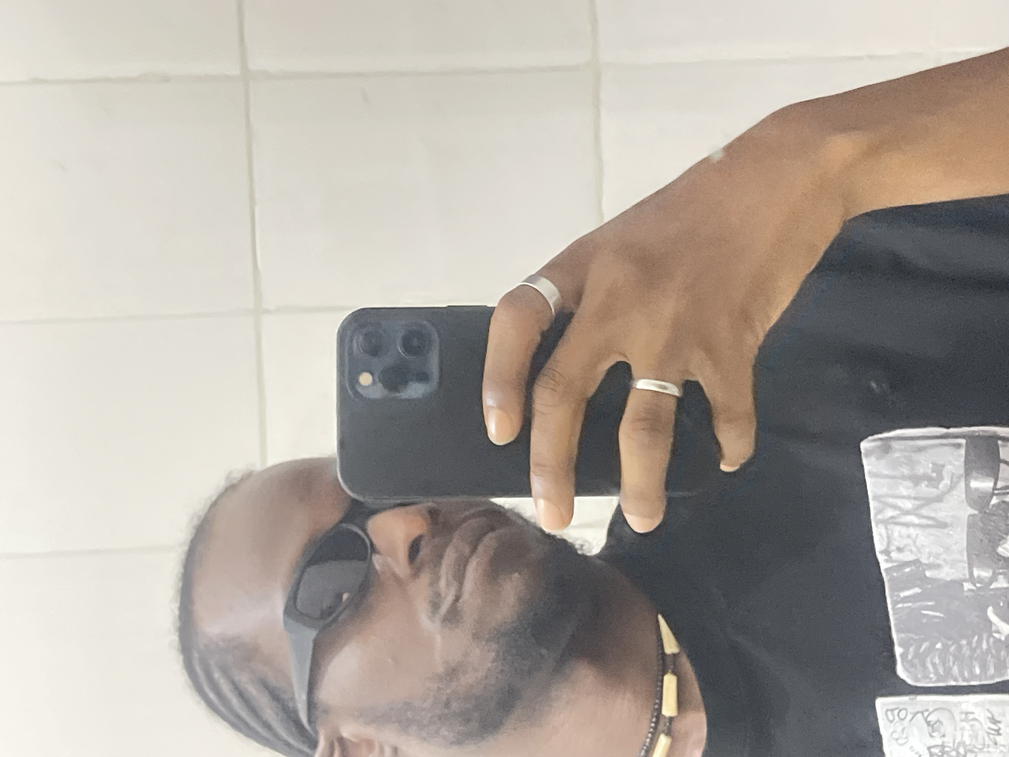 The image is a mirror selfie depicting a dark-skinned individual, appearing male, partially obscured by their black smartphone. The person wears black, angular sunglasses that cover their eyes, a black t-shirt with a monochrome graphic print, and a dark beaded necklace with light-colored, possibly bone or wood, rectangular beads. Their hair is styled in cornrows, and they have a short beard and mustache. The individual holds a black smartphone with a triple camera module (likely an iPhone) in their left hand, which is adorned with two plain silver-colored rings on the ring finger and the finger next to the pinky. The background is a light-colored, tiled wall, possibly white or off-white, with visible grout lines, indicative of an indoor setting. The specific urban location within Jos, Nigeria, is not discernible from the visual information presented in the image.