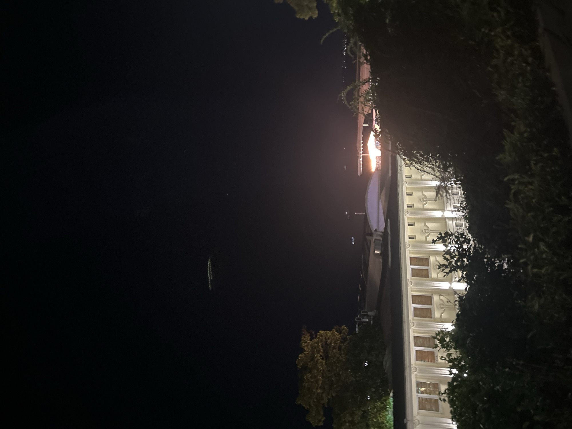 The image depicts a nighttime view of a coastal area in Varna, Bulgaria, likely overlooking the Black Sea. In the foreground, dark foliage partially obscures a building with a white, classical facade. The building is illuminated by several bright vertical lights positioned between its windows, which appear to be covered with wooden shutters. The building's architectural style suggests it could be a historical structure or a hotel.
In the middle ground, several other buildings and structures are visible, some with illuminated windows. A bridge or elevated walkway crosses over a body of water in the distance, with lights reflecting on its surface. Further out, a cluster of lights suggests a distant cityscape or port activity.
The sky above is dark, indicating nighttime. A peculiar rectangular object with a series of illuminated elements is suspended in the upper left portion of the sky, its nature unclear. The overall scene conveys a serene, albeit slightly mysterious, atmosphere of a city at night by the sea.