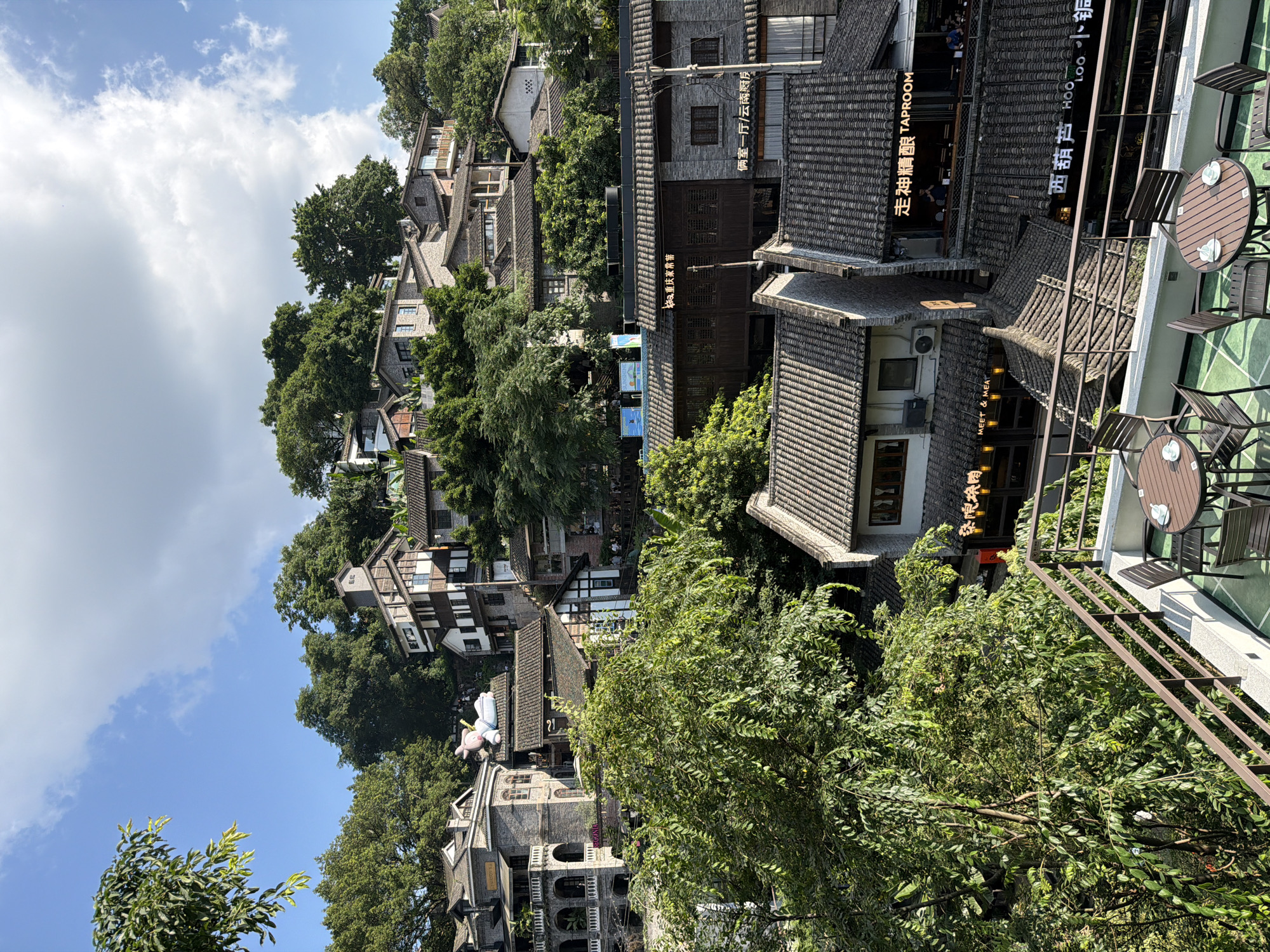 The image depicts a dense urban landscape with traditional Chinese architecture, situated on a hillside. The buildings are predominantly made of stone and tile, with multi-story structures cascading down the slope. Lush green trees are interspersed among the buildings, indicating a natural element within the urban setting.
In the foreground, a balcony with outdoor seating furniture, including tables and chairs, is visible. This balcony appears to overlook the cityscape. The tables are set with teacups, suggesting a cafe or restaurant setting.
The architecture itself is characterized by intricate details, such as arched windows, balconies with railings, and dark tiled roofs. Some buildings feature lighter colored facades with darker trim, while others are constructed entirely of stone. Signage with Chinese characters is visible on several buildings, indicating commercial establishments. One sign reads "TAPROOM" in English.
The sky above is a mix of blue and white clouds, suggesting a clear or partly cloudy day. The overall impression is one of a vibrant, densely populated area that harmoniously blends historical architecture with natural greenery.
Based on the distinctive architecture and style, this scene is consistent with the Hongyadong area in Nan'an District, Chongqing, China. The terraced buildings, steep hillside, and traditional design elements are characteristic of this popular tourist destination.