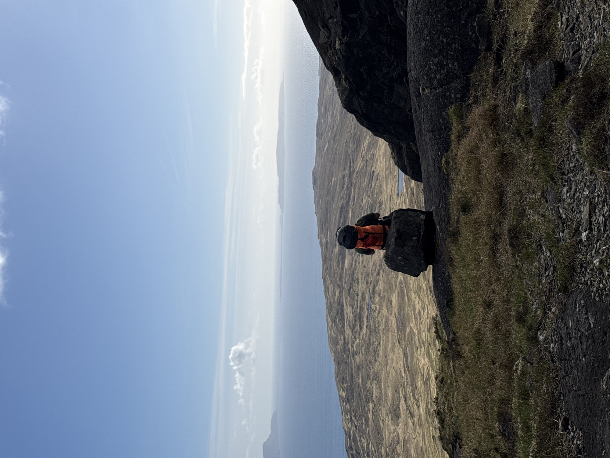 A person wearing an orange jacket and helmet is perched on a rock outcropping on a cliffside. The background reveals a view of a body of water, possibly the ocean, with land visible in the distance under a blue sky with scattered clouds. Vegetation such as grass and shrubs can be seen growing on the rocky terrain near the edge of the cliff. The location appears to be a mountainous region.