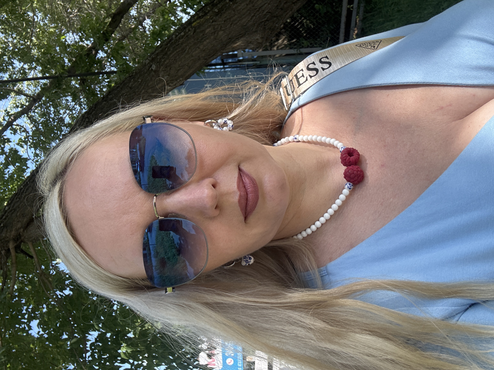 The image shows a close-up of a woman with blonde hair and fair skin, wearing sunglasses, earrings, and a light blue dress. She is also wearing a necklace with white beads and two red raspberry-shaped charms. A portion of a beige strap with the word "GUESS" printed on it is visible over her shoulder. The background consists of trees and foliage, suggesting an outdoor setting. There is no specific indication of the city of Komsomolsk-on-Amur, Russia within the image content. The woman is looking towards the camera, with a slight smile. The lighting suggests it is daytime, possibly sunny, given the reflections in the sunglasses and the shadows.