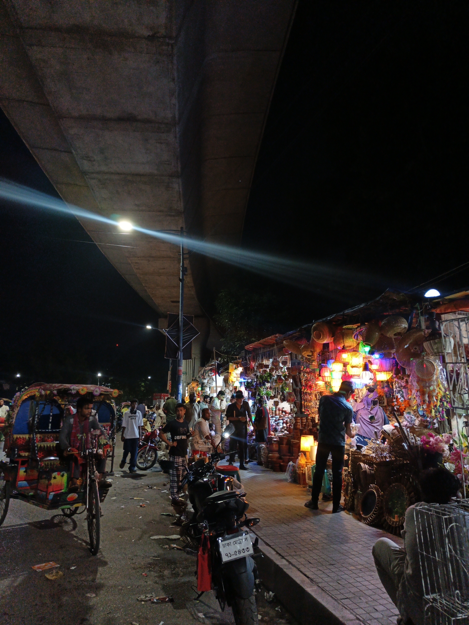 The image depicts a vibrant night market scene, possibly in South Asia, given the presence of a colorful cycle rickshaw. Underneath a large concrete overpass, a bustling marketplace is in full swing. The scene is illuminated by a street lamp that casts a bright beam of light, and numerous colorful lights from the stalls themselves, creating a lively and festive atmosphere.
Numerous people are visible, browsing the stalls, walking, and interacting. There are vendors displaying a variety of goods, including intricately woven baskets and lanterns that are brightly lit, dreamcatchers with colorful feathers, and various other handicraft items. A cycle rickshaw, decorated with floral patterns and vibrant colors, is positioned in the foreground, hinting at local transportation methods. Several motorcycles are also parked around the area.
The time of day is clearly night, with the dark sky providing a stark contrast to the illuminated market. The ground is paved with bricks, and there is some scattered litter, typical of a busy public space. The overall mood is one of activity and commerce, with the diverse array of lights and people suggesting a lively evening market.
Visible text includes a license plate on one of the motorcycles, which appears to be in Bengali script. The text reads "ঢাকা মেট্রো ল" followed by a series of numbers and Bengali characters. Another piece of text on a white t-shirt worn by a man in the crowd reads "DREAMS WORLD".