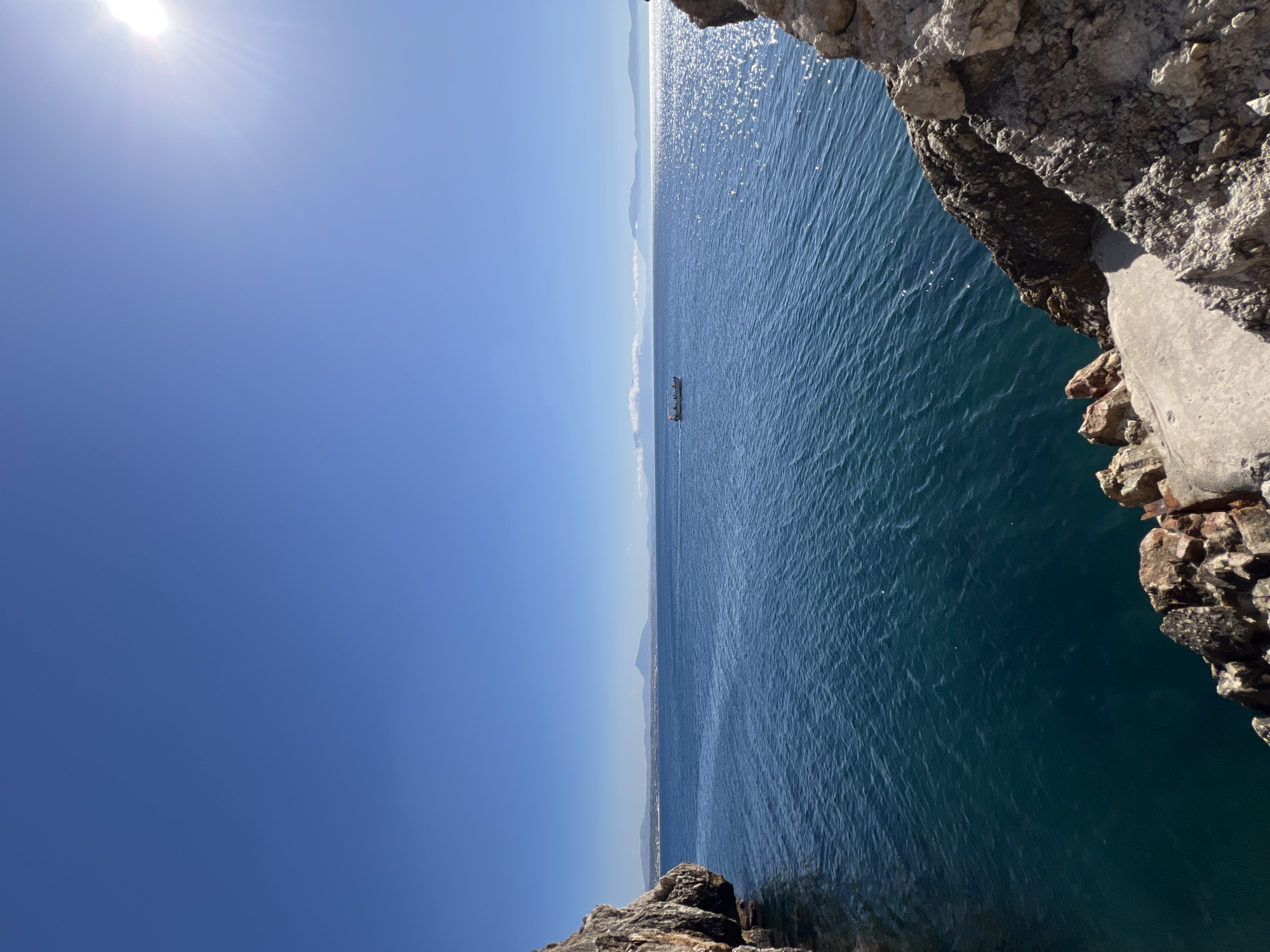 This media file captures a panoramic view of the sea and sky, likely from a high vantage point such as a cliff or rocky outcrop, given the presence of large rocks in the foreground. The scene is bathed in bright sunlight, suggesting a clear day, possibly late morning or early afternoon.
The vast expanse of the ocean dominates the right side of the frame. The water is a deep blue, with a shimmering surface due to the sun's glare, creating a textured appearance of ripples. In the distance, a small vessel with several figures on board can be seen sailing on the water. The horizon line is clearly defined, separating the sea from a hazy, mountainous landscape in the far distance. The sky is a vibrant, cloudless blue, with the sun positioned in the upper left corner, casting its bright rays.
The rocky foreground, on the right and bottom edges of the image, is rough and textured, hinting at a natural, untamed environment. The presence of rocks suggests a coastal setting.
The overall mood of the image is serene and expansive, emphasizing the beauty of the natural landscape. The clear weather and bright sunlight contribute to a feeling of openness and tranquility. No text is visible in the media file. Given the location context of Terrasini, Italy, the scene is likely a view of the Mediterranean Sea.