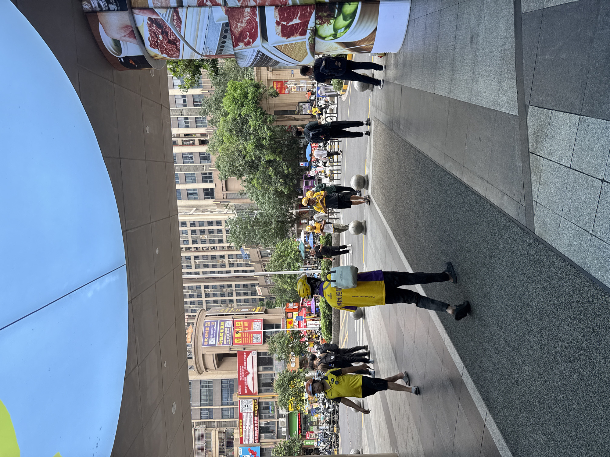 This is a street-level, full-shot photograph taken from a slightly elevated angle in Haizhu District, China. The scene depicts a pedestrian area with a curved canopy overhead that is partially visible. The foreground is dominated by a walkway with textured grey paving and a dark grey speckled strip.
Several people are present in the image. In the mid-ground, two individuals in yellow shirts and helmets are walking on the paved area. The person on the left wears a yellow shirt and dark shorts, while the person on the right wears a yellow shirt with purple accents and black pants, carrying a light blue tote bag over their shoulder. Further back, a group of individuals, some in yellow shirts and helmets, are walking or standing. One person in the background is wearing black clothing and a backpack.
The environment includes buildings with multiple stories in the background, some featuring balconies and windows. Trees and greenery are visible between the buildings. A large, curved pillar on the right side of the image features a collage of images, appearing to be advertisements or decorations related to food, specifically raw meat. There are bollards along the walkway, and a line of parked motorcycles can be seen in the distance on the left. The overall impression is a busy urban commercial or public space.