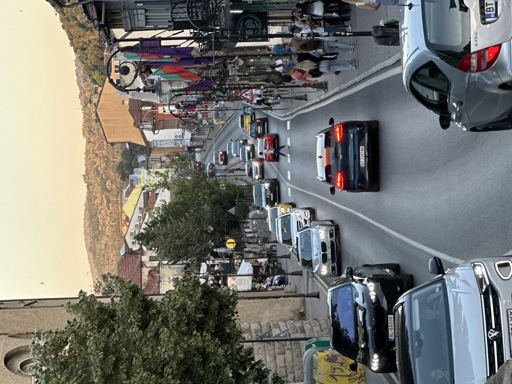 This is a vertical, medium-distance shot of a busy street in Veliko Tarnovo, Bulgaria, during the golden hour. The street slopes downwards and is lined with buildings and trees. Several cars are in traffic, including a black BMW X6 with license plate BL 8888B in the foreground, a silver Volkswagen T-Roc behind it, and a dark Kia with license plate BT 3141 MA further down the road. Pedestrians are visible on the sidewalks, some interacting with each other and others entering or exiting shops. Bulgarian flags are displayed on poles along the street, indicating a national celebration or event. The background shows a hillside covered with trees and buildings, bathed in warm sunlight. The scene conveys a sense of vibrant urban life in the historic city.