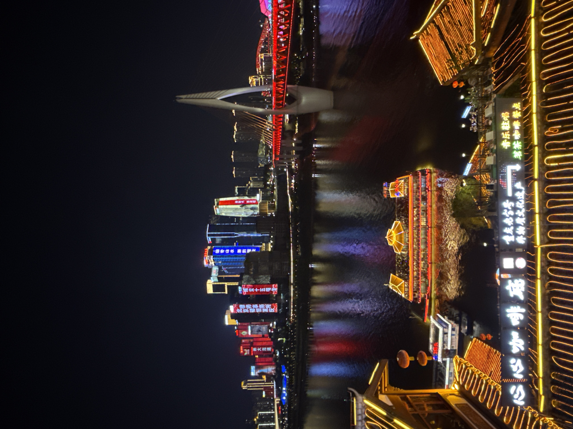The image captures a nighttime view of the Yuzhong District in China, showcasing a vibrant cityscape along a waterfront. In the foreground, traditional Chinese-style buildings with illuminated roofs and railings are visible, casting a warm glow. A large, multi-story boat with illuminated windows and balconies is docked on the water. Further in the distance, a modern bridge with a distinctive white, arching support structure spans the river, its red illuminated deck adding a dramatic contrast to the night sky. The background features a cluster of modern skyscrapers, adorned with colorful neon lights and digital advertisements. The river reflects the bright lights of the city and the bridge, creating a shimmering effect on the water's surface. The sky is dark, emphasizing the brilliance of the artificial lighting.