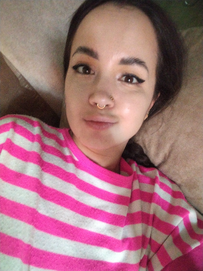 A young woman lies against tan cushions wearing a pink and white striped sweater. She faces the camera with dark eyes and arched eyebrows. Her makeup includes winged eyeliner and defined brows. She sports a gold nose ring and what appears to be a small gold earring. Her hair is dark and pulled back. The lighting is soft and natural, highlighting her skin tone.