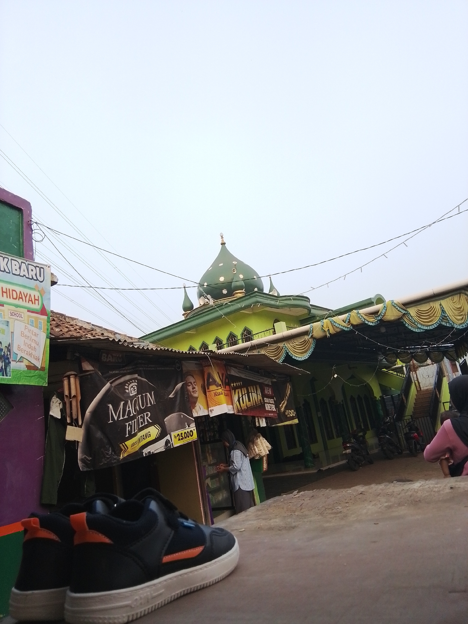 The image shows a street scene in Bencongan, Indonesia, featuring a mosque with a green dome. In the foreground, a pair of black and orange sneakers are placed on the ground. Behind the mosque, there is a building with a tiled roof and awnings. Several posters are visible, including one advertising "MAGUM FIER" for Rp 25,000. A few people are present in the background, some wearing headscarves. Motorcycles are parked near the mosque. The sky is overcast. The overall scene suggests a market or community gathering area outside a religious building.