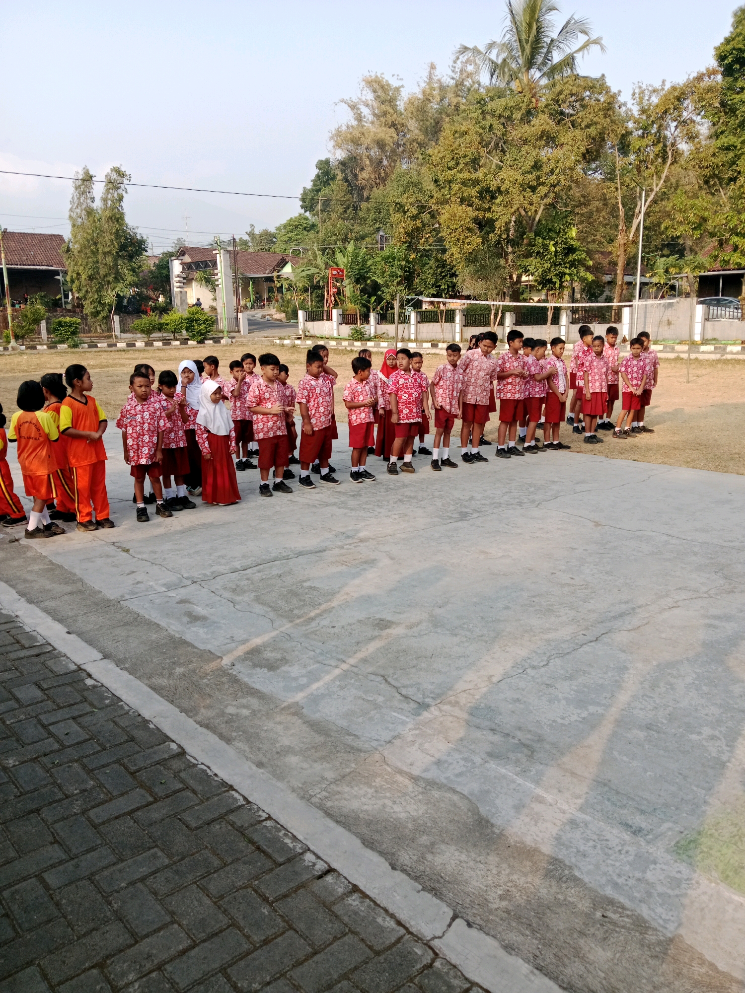 A group of children in school uniforms stand in a line on a paved surface outdoors, in front of a grassy area and buildings in Salatiga, Indonesia. The children are diverse in age and gender, with some wearing white hijabs. They appear to be in formation, possibly for a school activity or assembly. The background shows trees, houses, and a fence. The ground is made of concrete with some cracks, and a section of dark paving stones is visible on the left. The lighting suggests it is daytime, likely morning or afternoon.