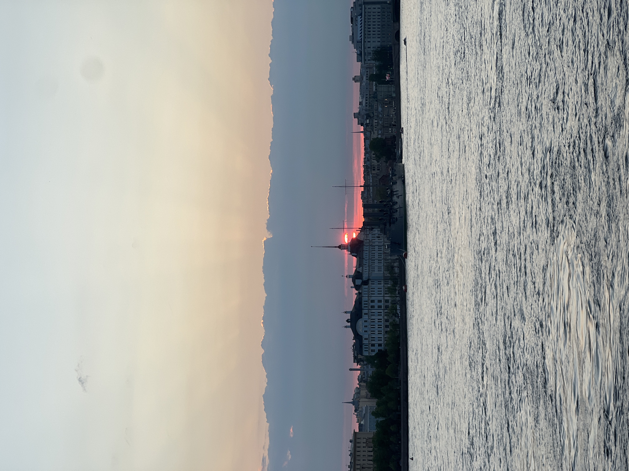 The image captures a sunset over the Neva River in Saint Petersburg, Russia. The sun is partially obscured by the horizon and clouds, creating a red glow. A cityscape is visible on the riverbank, with buildings of varying heights and architectural styles. The Peter and Paul Cathedral is seen in the background and provides a landmark to the shot, along with other buildings. The Neva River reflects the sunset colors, showing ripples and movement of the water. The sky transitions from a pale white to orange near the horizon, blending into a darker blue above. The image is rotated 90 degrees.