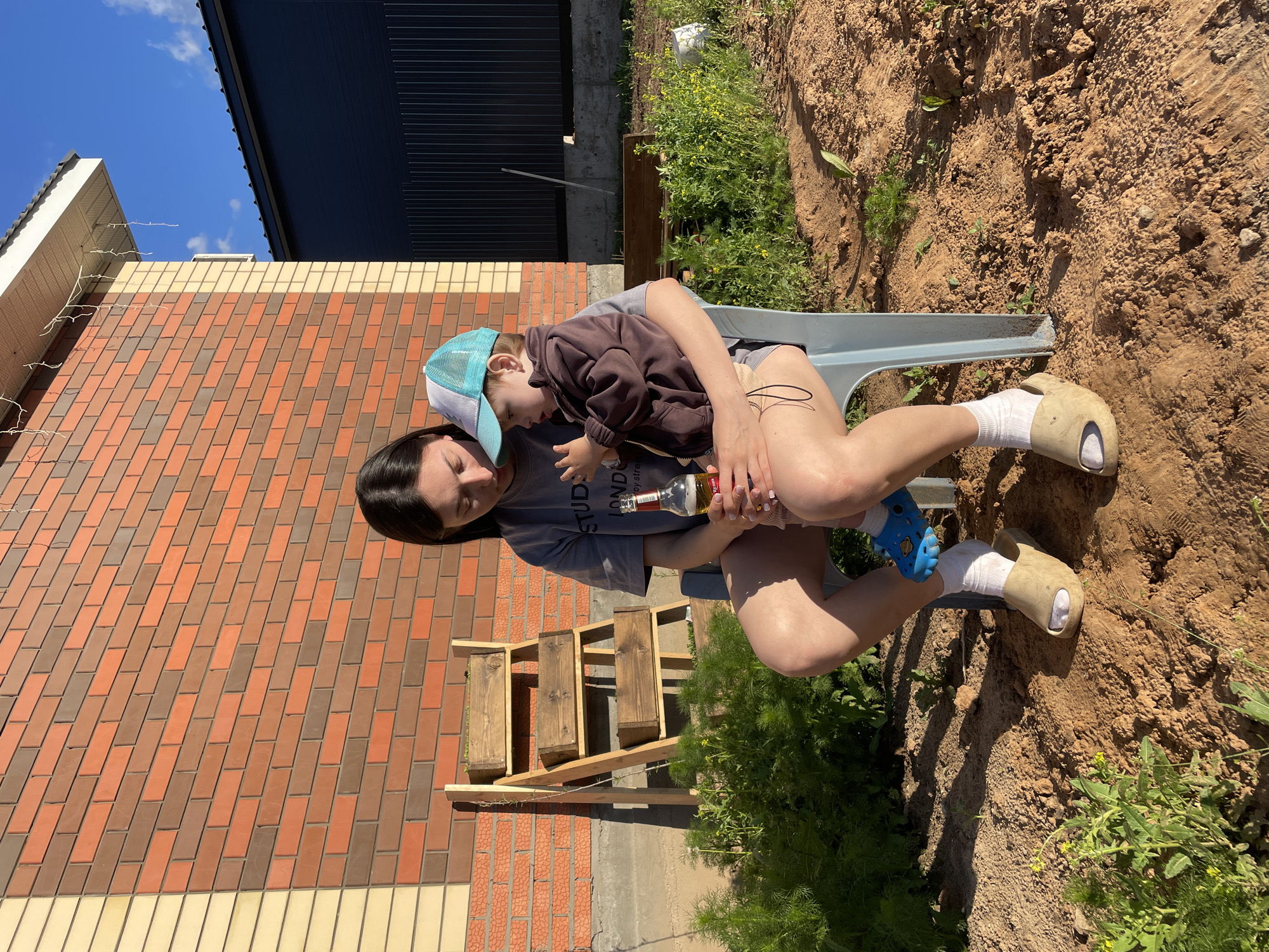 A woman sits outside in a backyard holding a toddler in her lap. The woman has long dark hair, wears a gray t-shirt and shorts. The toddler wears a brown sweatshirt, a blue baseball cap, and blue sandals. The woman sits in a light blue plastic chair. She is holding a glass bottle. There is a brick building behind them with a dark roof extension. Wooden planters are nearby. The ground is mostly dirt with some patches of green plants. The location may be Эстачи, Russia.