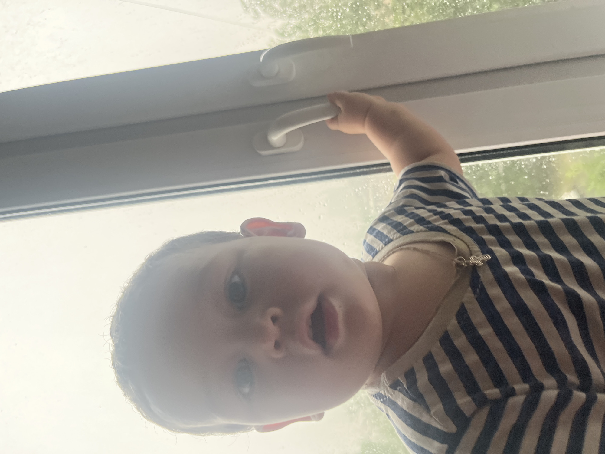 The image shows a close-up, low-angle, vertical shot of a baby reaching for a handle on a white window frame. The baby, with wide, surprised eyes, is wearing a blue and white striped shirt. The window is likely in a home. Outside the window, it appears to be raining, with raindrops visible on the glass, suggesting a cloudy or overcast day. The lighting is bright and diffused, indicating it is daytime. The baby's expression and grasp on the handle suggest curiosity and a desire to interact with the window mechanism. There is no visible text in the image. Given the location context of Chelyabinsk, Russia, this could be a common domestic scene during inclement weather.