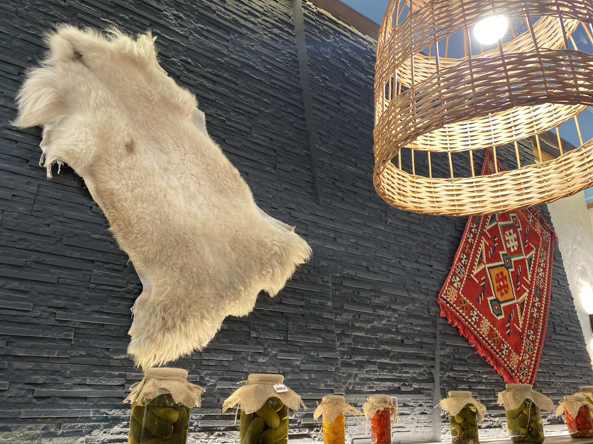 This image captures a rustic and culturally-inspired interior scene, likely within a restaurant or traditional shop in Kazan, Russia. No people or active events are depicted.
The focal point on the left is a light-colored, fluffy fur pelt, possibly sheepskin, hanging vertically against a dark gray wall. This wall is composed of narrow, horizontally stacked stone or tile slats, creating a textured backdrop.
On the right side of the image, a large, cylindrical lampshade made of woven light-colored wicker hangs from the ceiling, illuminating the scene with a bright light bulb visible inside. Below the lampshade, a vibrant red rug or tapestry with geometric patterns in yellow, green, and white is suspended diagonally on the textured wall, adding a splash of color and traditional artistry.
Along the bottom of the image, a row of glass jars filled with various pickled vegetables—predominantly green cucumbers, but also some yellow and red items resembling pickled peppers or tomatoes—are arranged on a shelf or counter. Each jar is topped with a small piece of burlap tied with string, giving them a homespun appearance. Small white tags or labels are visible on some of these burlap covers, though the text on them is indistinct and unreadable.
The overall atmosphere suggests warmth and a connection to traditional folk elements, characteristic of the region, emphasizing natural materials and preserved foods. The lighting is artificial, suitable for an indoor setting, and there are no discernible external weather conditions.