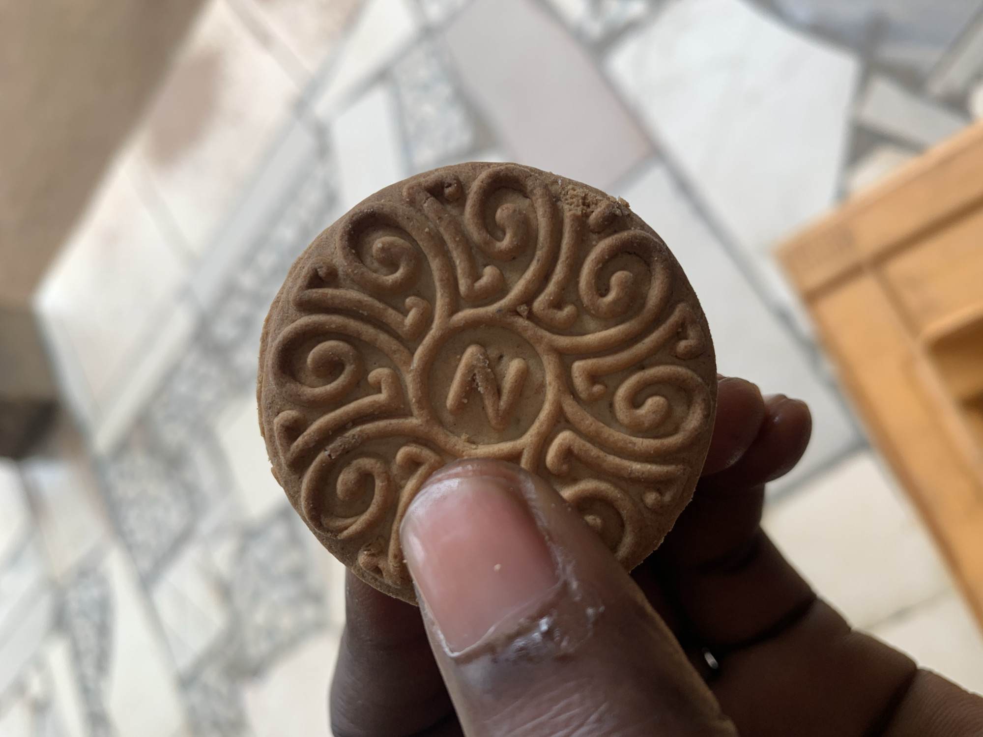 A hand holds a patterned, round biscuit featuring a prominent letter "N" in its center. The biscuit is light brown with swirling designs. The hand has dark skin and a slightly shiny appearance. The background is a blurred combination of patterned tiles and a light brown wooden surface. The photo's location is indeterminable based on the visual information provided, so I cannot confirm whether this image was taken in New Stadium, Nigeria.