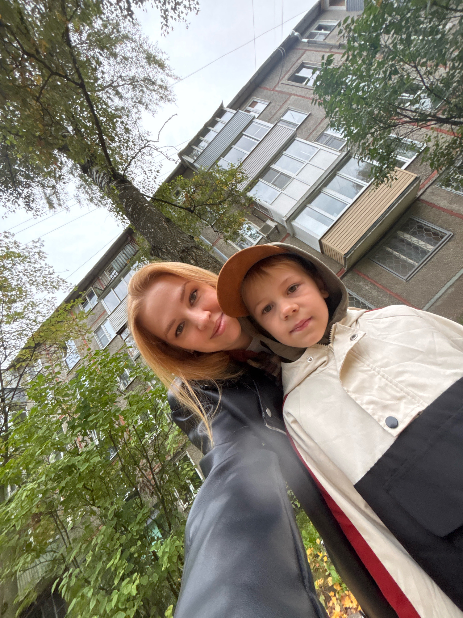 In a selfie taken outdoors in front of a Soviet-era apartment building in Rusanovo, Russia, a woman with blonde hair and a young boy are prominently featured. The woman, positioned on the left, is wearing a black leather jacket and smiling at the camera. The boy, on the right, is looking directly at the camera and wearing a hooded jacket with beige, black, and red accents. He also wears a brown cap. The apartment building, with its grey facade and numerous windows, serves as the backdrop. Lush green trees with some autumn leaves are visible, suggesting the season. The sky is overcast.