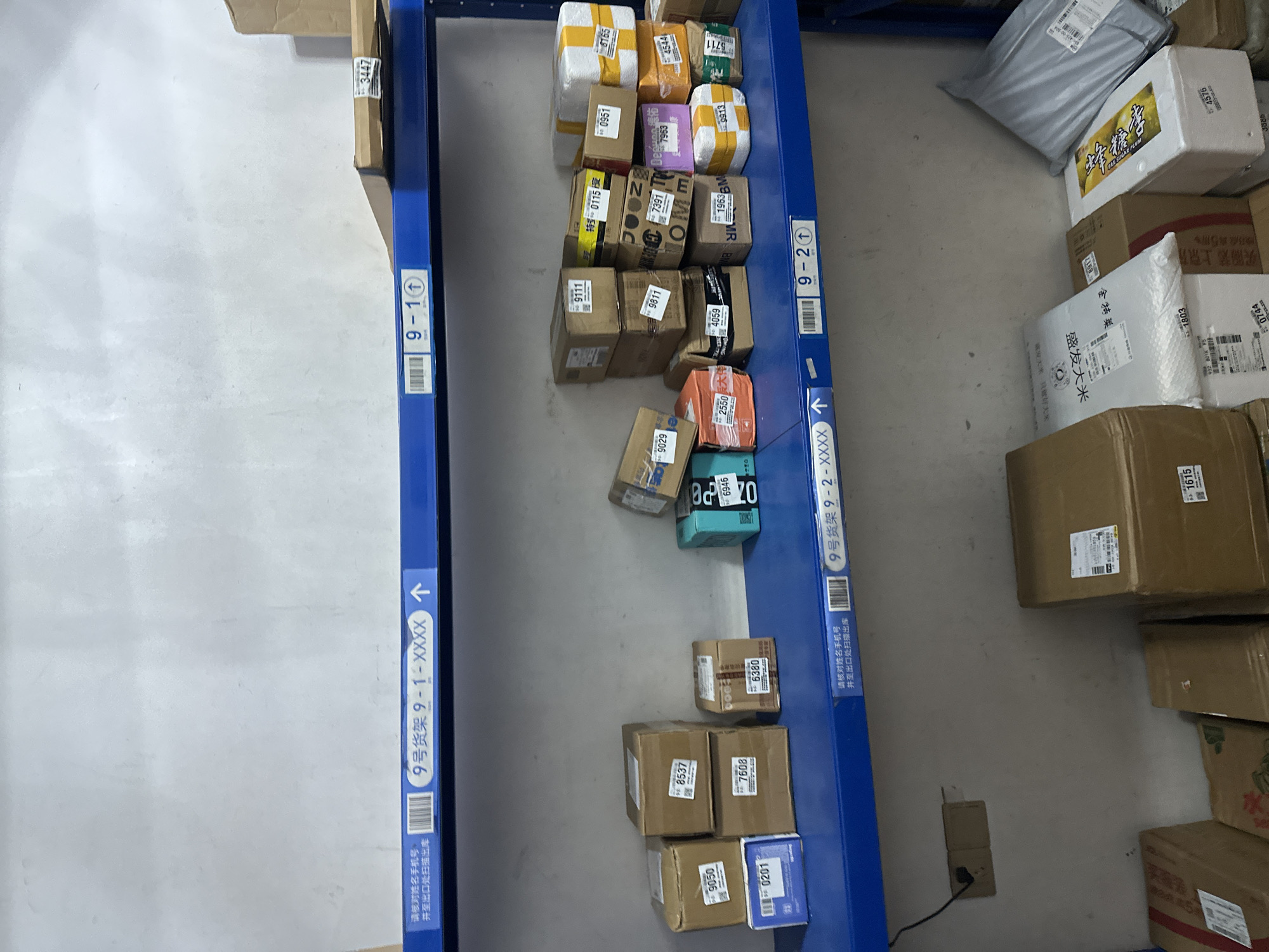The image depicts a storage shelf in a warehouse, likely in the Chongqing High-tech Zone, China. The shelves are blue metal, and multiple cardboard boxes are stacked on them. Some boxes have labels with numbers, such as "9050", "8537", "7608", "0201", "6380", "9029", "2550", "4059", "0111", "9811", "7391", "1963BM", "7963", "0951", "3447", "0115", "8165", "4544", "3711", "9913", and "4576". There are also some labels in Chinese characters. In the lower part of the image, more boxes and packages are visible, including some white bags with Chinese text. A black cable is plugged into an outlet on the wall behind the shelves. The overall environment appears to be an organized storage facility.